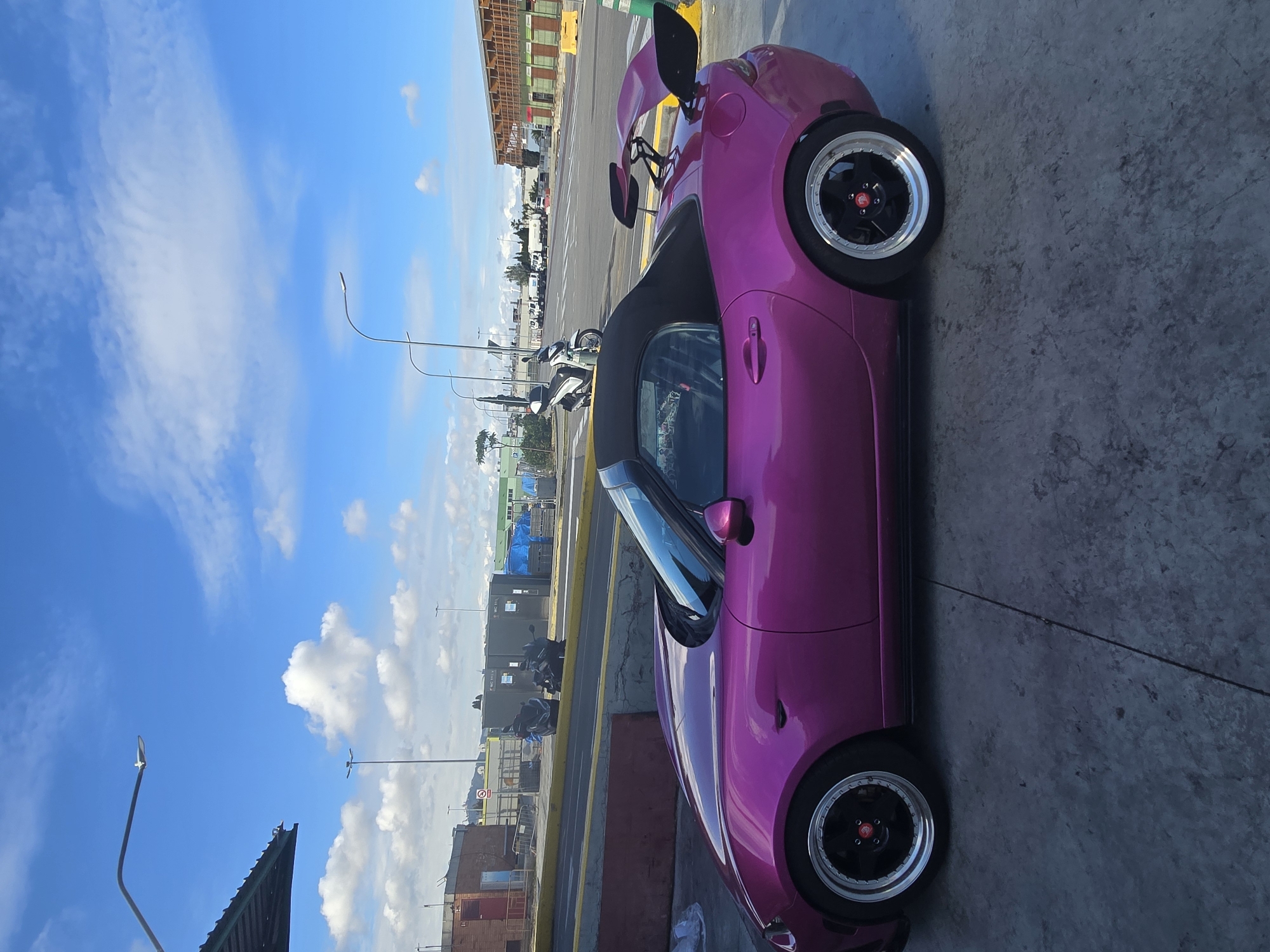 A magenta sports car is parked on concrete. The car has a spoiler. A black top is visible. There are buildings, light posts, and other cars in the background, as well as a blue sky with clouds. The setting appears to be urban. Based on the architectural style of the buildings, the location is possibly in Barcelona, Spain.