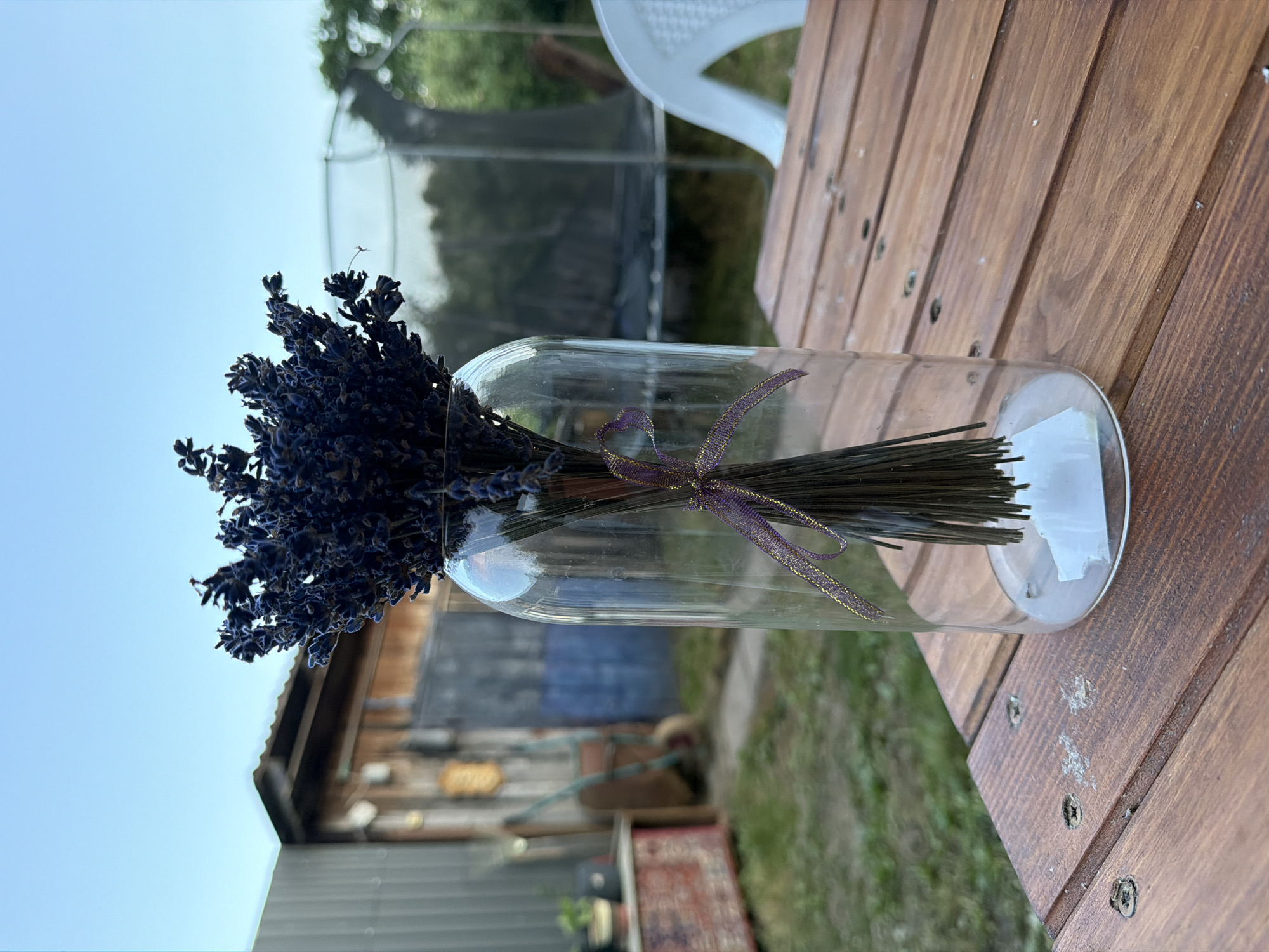 The image depicts a bouquet of dried lavender in a clear glass vase, placed on a wooden table outdoors. The lavender is tied with a purple and gold ribbon. In the background, there is a rustic wooden shed with a corrugated metal roof, a trampoline, and a white plastic chair. The scene appears to be in a rural or backyard setting. The sky is clear and pale blue.