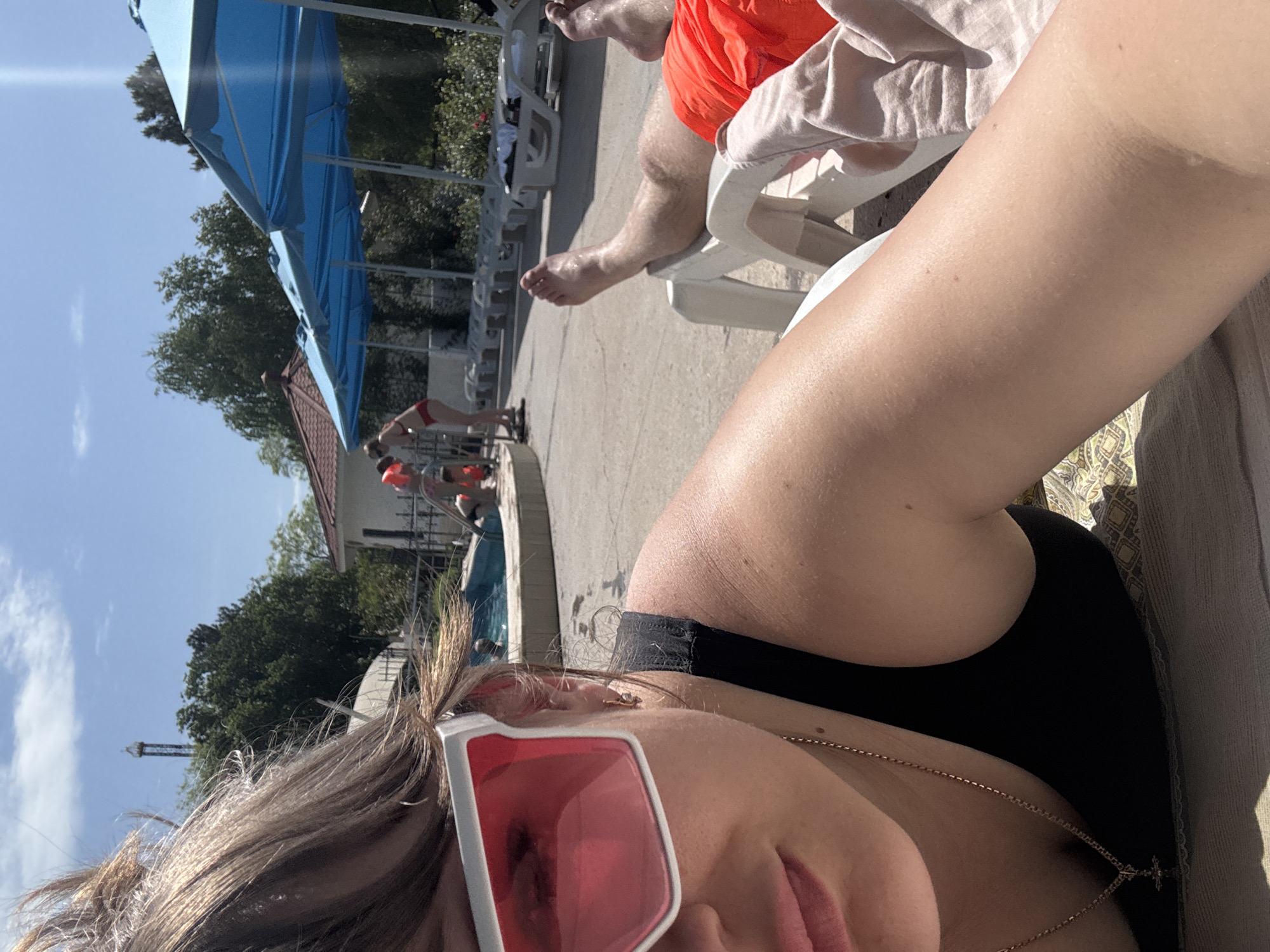 A partially-visible person in a black swimsuit and rose-colored glasses is photographed near a swimming pool. In the background, another person with orange shorts sits on a white chair. Blue canopies, trees, and buildings are also visible. This appears to be a poolside scene, possibly in the city of Yevpatoriya, Ukraine.
