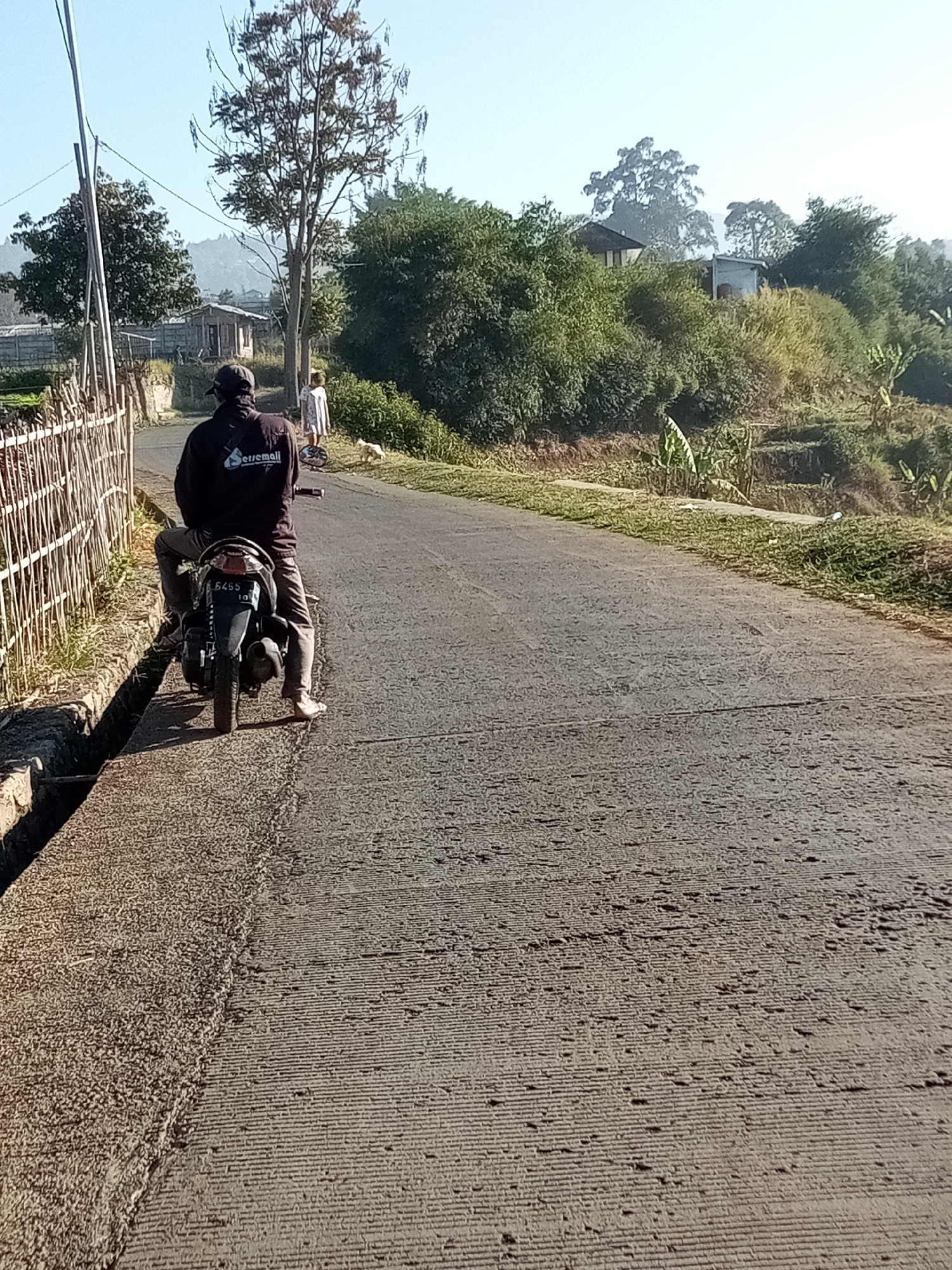 In Cihanjuang, Indonesia, on a paved road under a clear sky, a person wearing a dark jacket and a cap is seated on a motorcycle. The motorcycle's license plate reads "6455 10". To the left of the road, a wooden fence runs alongside a concrete drain. In the background, there are several houses and lush green trees. A young child in a white dress and a small dog are visible further down the road. The environment suggests a rural or semi-rural setting with agricultural terraces visible on the hillside.