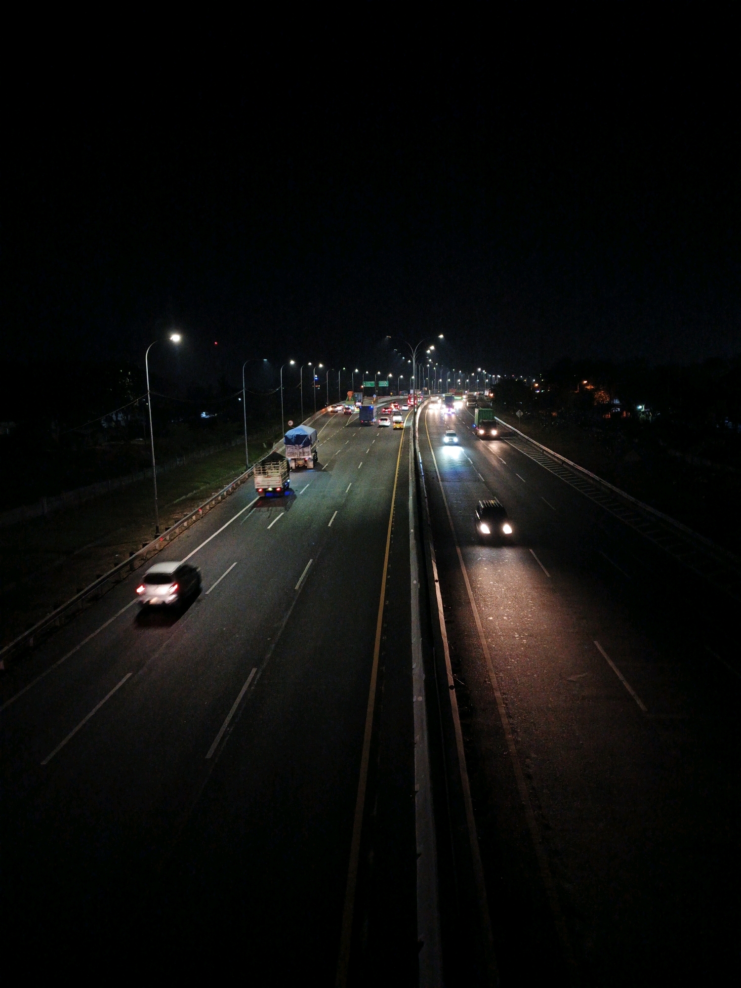 The image captures a nighttime scene on a highway, likely a toll road, in Kludan, Indonesia. The highway stretches into the darkness, illuminated by rows of streetlights and the headlights of passing vehicles. Multiple cars and trucks are visible, their lights creating streaks on the wet-looking asphalt. On the left side of the highway, there is a grassy embankment and a guardrail. The right side of the highway features a more substantial barrier and a darker, more vegetated area, suggesting it might be the edge of a forest or undeveloped land. The overall impression is one of constant motion and the stark contrast between artificial light and the deep night sky.