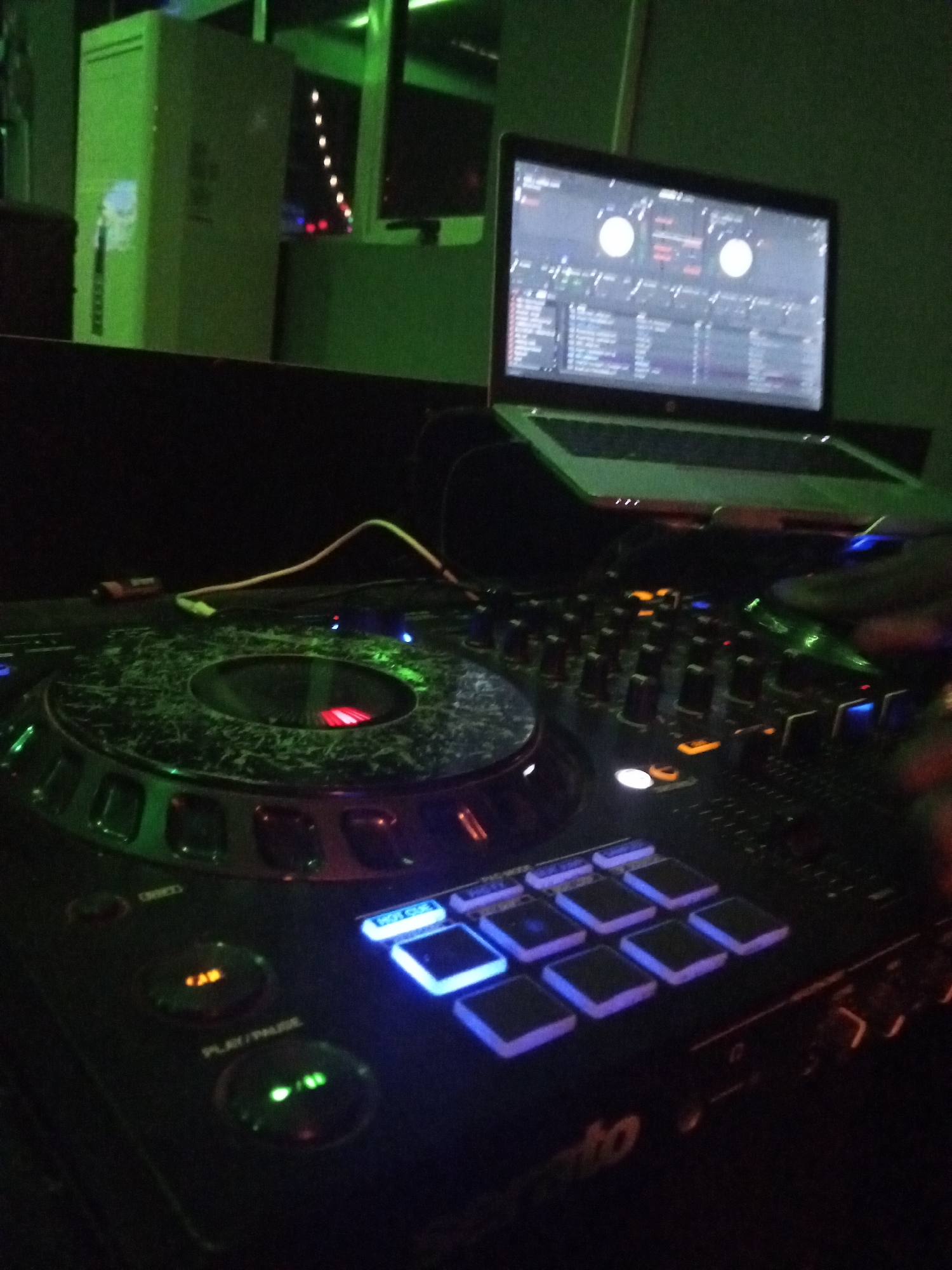 The image shows a DJ setup in a dimly lit environment, likely indoors. In the foreground is a DJ controller with illuminated buttons and knobs. A laptop displaying DJ software rests behind the controller. A hand is partially visible to the right. The background reveals a dark space with a green-lit box and possibly a window with faint lights. The location is unknown and no specifics about the city of Lokoja, Nigeria, can be inferred from the image.