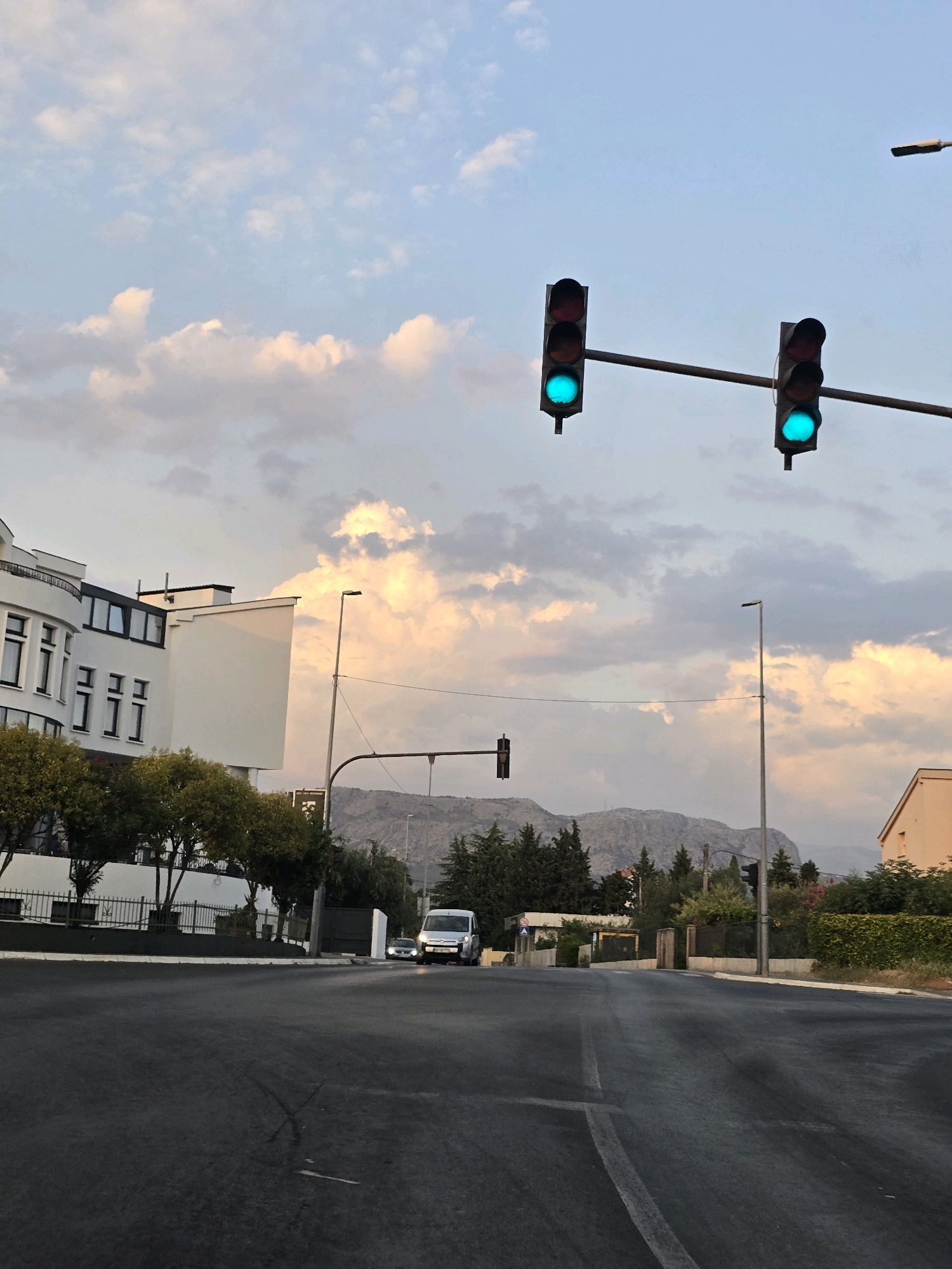 The image captures a street scene in Podgorica, Montenegro, likely during the late afternoon or early evening, given the soft, diffused light. Two sets of traffic lights are prominently displayed, both showing a green signal. The foreground is dominated by a dark asphalt road with white lane markings. To the left, a modern white building with multiple windows and balconies is visible, partially obscured by trees. Further down the street, a white van and a smaller car are approaching. The background features a dense line of trees and a mountainous landscape under a sky filled with soft, pastel-colored clouds. The overall atmosphere is tranquil and urban.