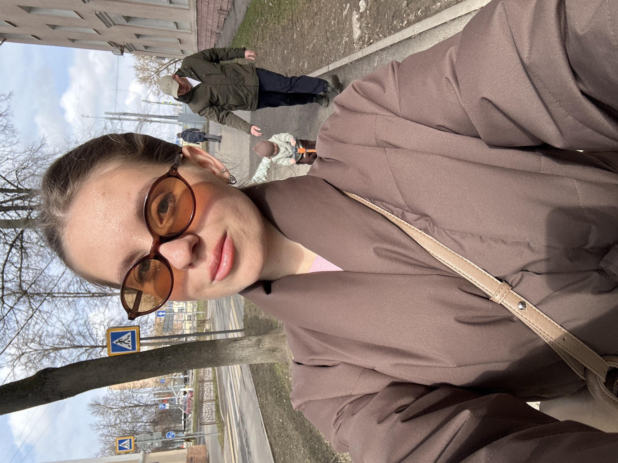 The image depicts a selfie taken outdoors, likely in Klin, Russia, judging by the Cyrillic characters on the road signs. The main subject is a fair-skinned person with light brown hair pulled back, wearing brown-tinted glasses and a brown jacket. A tan-colored purse strap is visible across their body. In the background, slightly out of focus, are several pedestrians: a person in a green jacket and light-colored cap, a person in dark clothing, and a child on a push toy. The setting appears to be a sidewalk alongside a street, with trees visible, suggesting an urban or suburban area. There are road signs with blue backgrounds bearing a symbol that appears to be two people walking and a pedestrian crosswalk. The buildings are of modest size and construction. It is daytime, with diffused sunlight creating soft shadows.