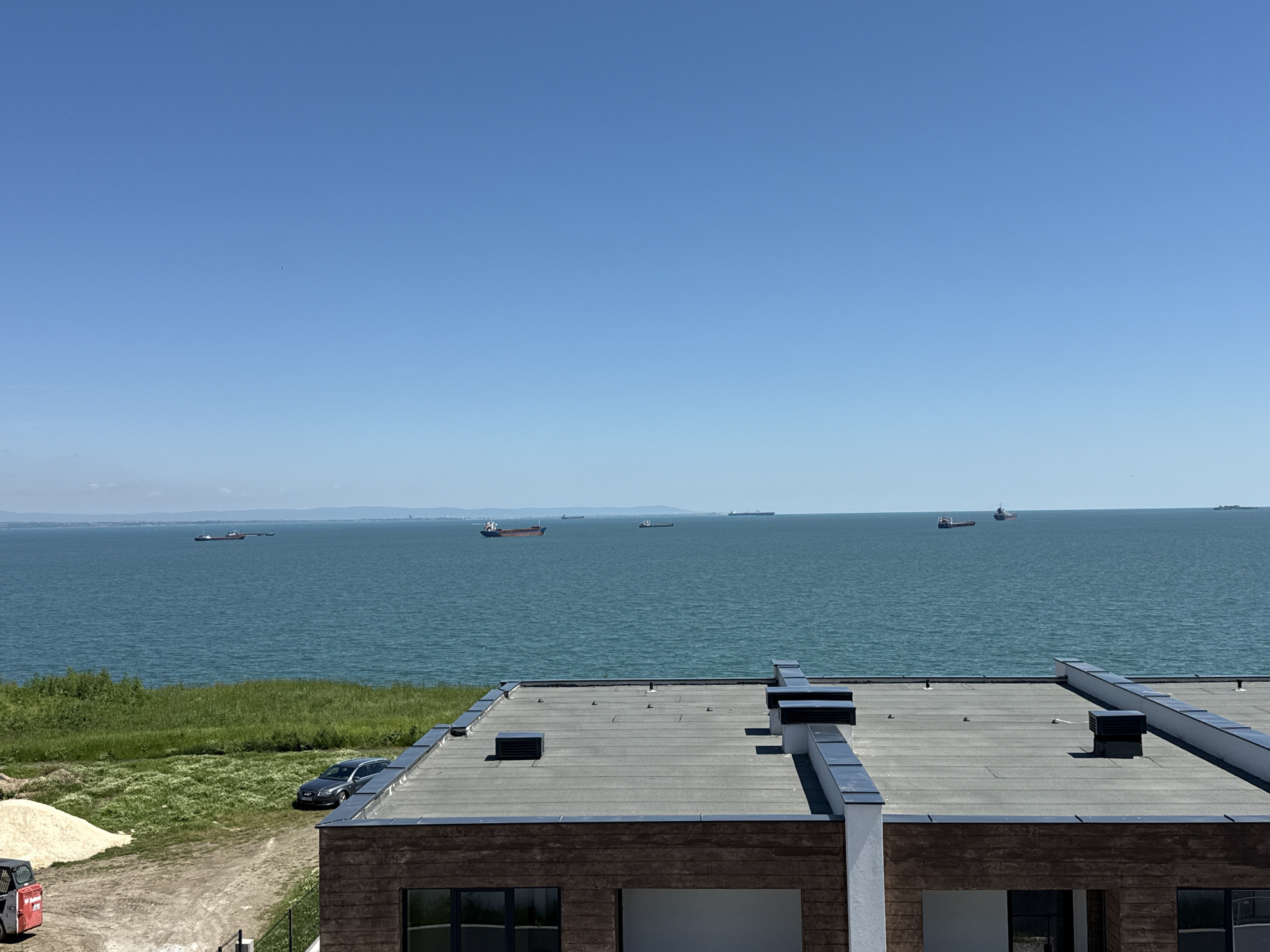 The image presents a cityscape with a view of the sea, likely taken from an elevated position in or near the city of Burgas, Bulgaria, given the geographical context of the Black Sea. The sky is clear and blue. Several ships are scattered across the water's horizon.
In the foreground, the flat roof of a building with a gray surface and dark metallic accents is visible. The building appears to be modern in design, with a brick-like exterior and dark-framed windows. Next to the house is a green area with grass, and a silver/grey car is parked. Beside the car, there is a sand pile and a small red excavator.