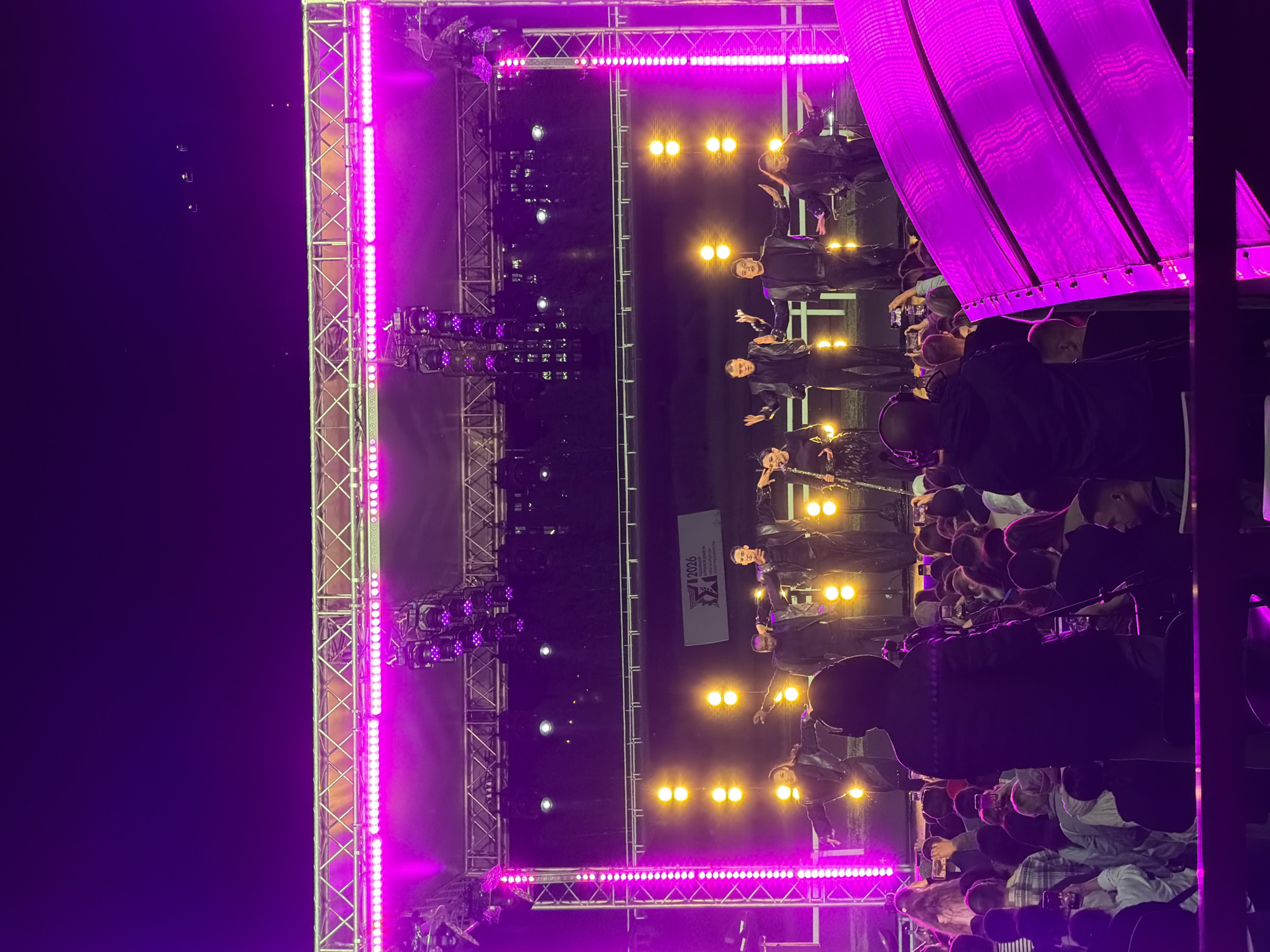 An outdoor concert stage is illuminated with pink and yellow lights at night. A group of performers in dark, matching outfits stand on the stage, some with their arms outstretched. A large crowd of people is gathered in front of the stage, many holding up their phones to take pictures or record videos. The audience is silhouetted against the bright stage lights, making it difficult to discern individual features. The overall scene suggests a lively and energetic event.