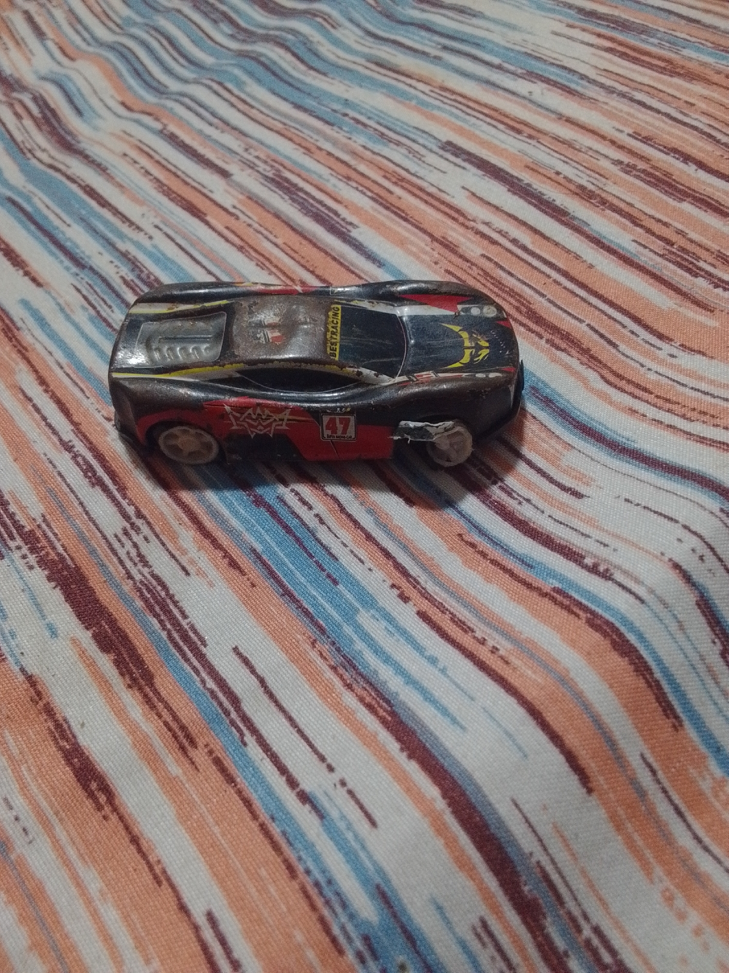 A close-up, high-angle shot shows a weathered, black toy race car with red and yellow accents resting on a striped fabric. The fabric is patterned with diagonal lines of white, light orange, and dark reddish-brown, with occasional thin blue stripes. The toy car is a low-profile, futuristic design with a transparent windshield.
Visible text on the car includes "BESTRACING" in yellow lettering along the top rear of the car. On the side of the car, a red rectangular decal with the number "47" is visible, and below it, smaller text reads "SUPER RACING CAR".
The car appears to be slightly worn, suggesting it has been played with. The lighting is even, indicating it might be an indoor setting or an overcast day. No people are visible in the frame. The overall scene is a simple still life of a toy car on a textured surface. The location context of La Tagua, Venezuela, does not provide specific visual cues within the image itself, but implies a domestic or casual setting.