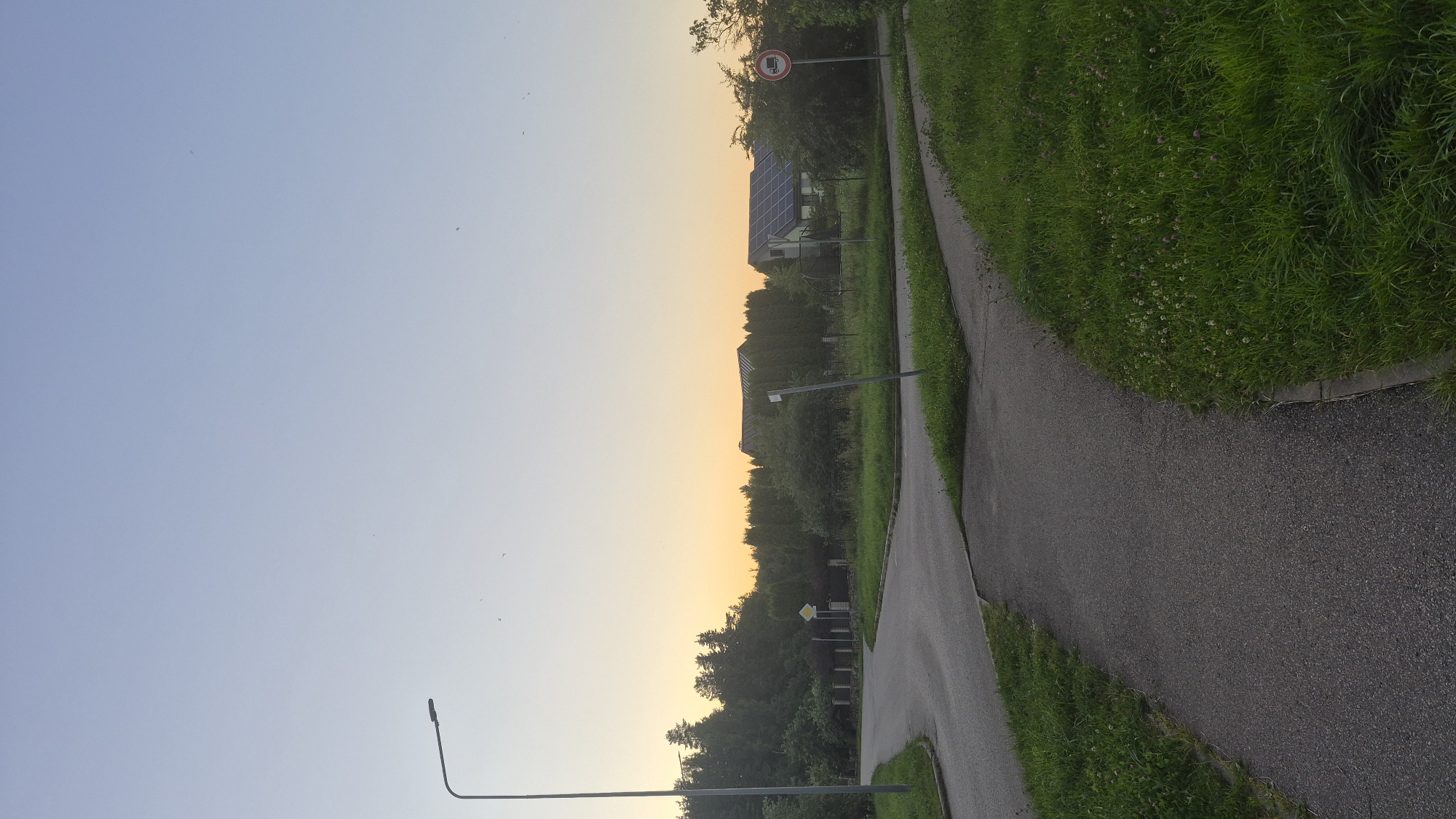 The image depicts a street scene in Krauja, Latvia, during the golden hour of sunset. A paved road curves into the frame from the left, with a separate asphalt footpath running parallel to it on the right. Both are bordered by lush green grass. In the background, several houses with solar panels on their roofs are visible, nestled among dense trees and hedges. A streetlamp stands tall on the left side of the road. Traffic signs are present, including a "No Trucks" sign on the right. The sky is a gradient of pale blue and soft orange, indicating the setting sun. No people or animals are present in the scene.