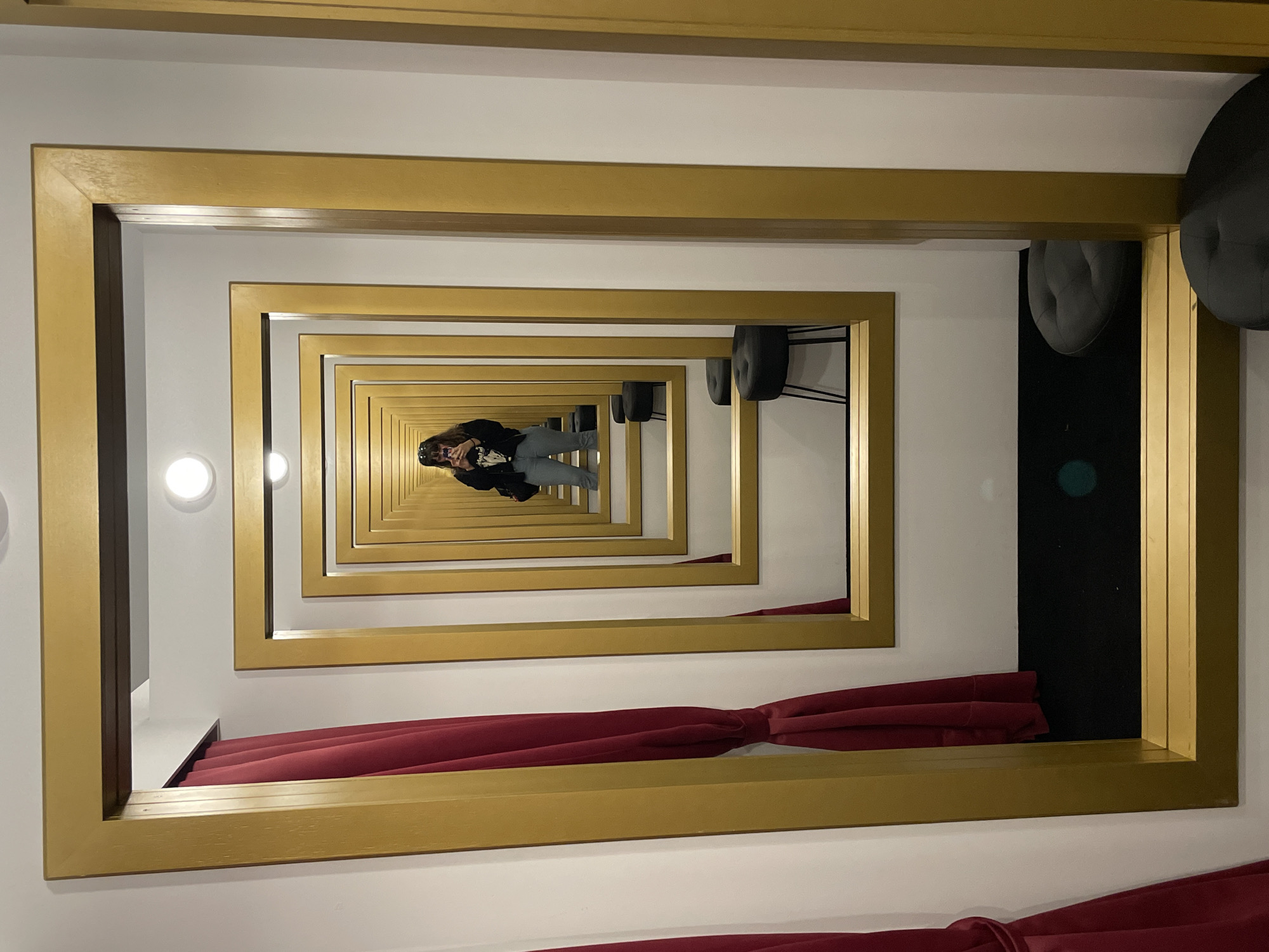 The image shows a hallway lined with diminishing golden frames creating an infinity mirror effect. A person is visible at the center of the hallway, reflected in the mirrors, wearing a black jacket and light-colored pants, holding a mobile phone, seemingly taking a picture of themselves. A red curtain is visible at the bottom and a couple of gray chairs on the right side. Based on the artistic installation, this image is likely taken at the Espai Mesmerism Gallery in Barcelona, Spain.