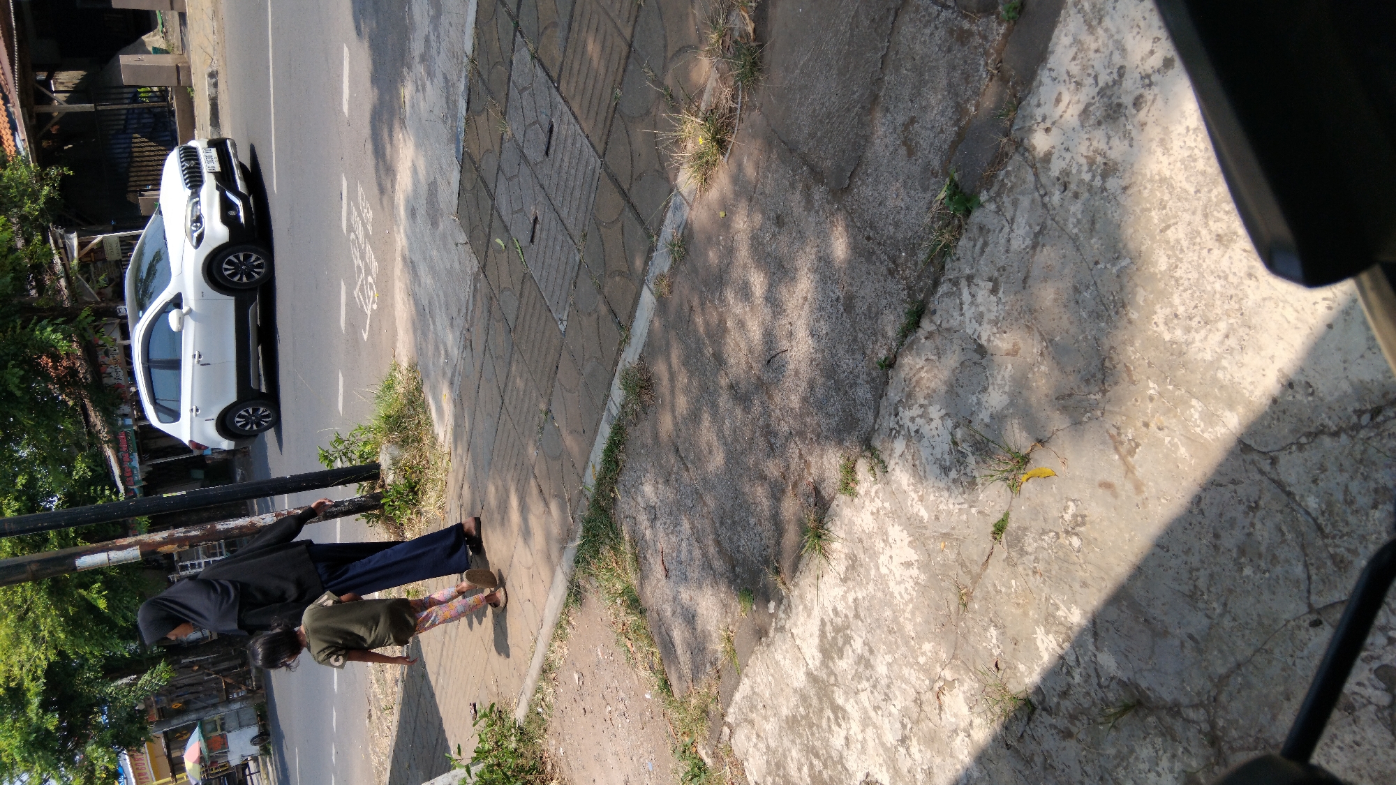 The image shows a woman in a black hijab and a child walking on a sidewalk next to a road in Proyonangg, Indonesia. The woman is wearing a long blue skirt. The child is wearing a green shirt and patterned leggings. A white SUV is driving on the road behind them. The sidewalk is made of concrete and pavers. There are trees and buildings in the background. The sun is shining, casting shadows on the sidewalk.