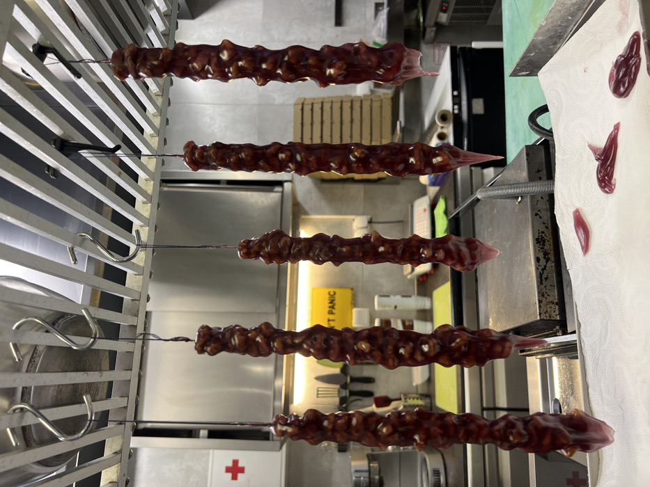 The image depicts a commercial kitchen in Budva, Montenegro, where five elongated food items, consistent with a type of confectionery like Churchkhela, are hanging vertically from S-hooks attached to a metal overhead rack for drying. These items are dark reddish-brown, textured, and appear to contain nuts encased in a fruit paste. Liquid is visibly dripping from the lower ends of some items.
The background shows a professional kitchen environment with stainless steel surfaces, including countertops and cabinet doors. On the left, a white square with a red cross, indicating a first aid station, is visible on a cabinet. A yellow rectangular sign with the text "DON'T PANIC" is affixed to a wall in the mid-ground. Other kitchen equipment includes a silver commercial mixer on the left, a green cutting mat on a counter, and various utensils on a magnetic strip. Further back, a stack of what appears to be cardboard boxes, possibly pizza boxes, is present. Commercial-grade appliances are visible in the deeper background. Below the hanging items, a white paper towel laid on a metal tray collects the reddish-purple liquid drips. Several small, irregular puddles and solidified pieces of this liquid are visible on the paper towel. No individuals are present in the scene.