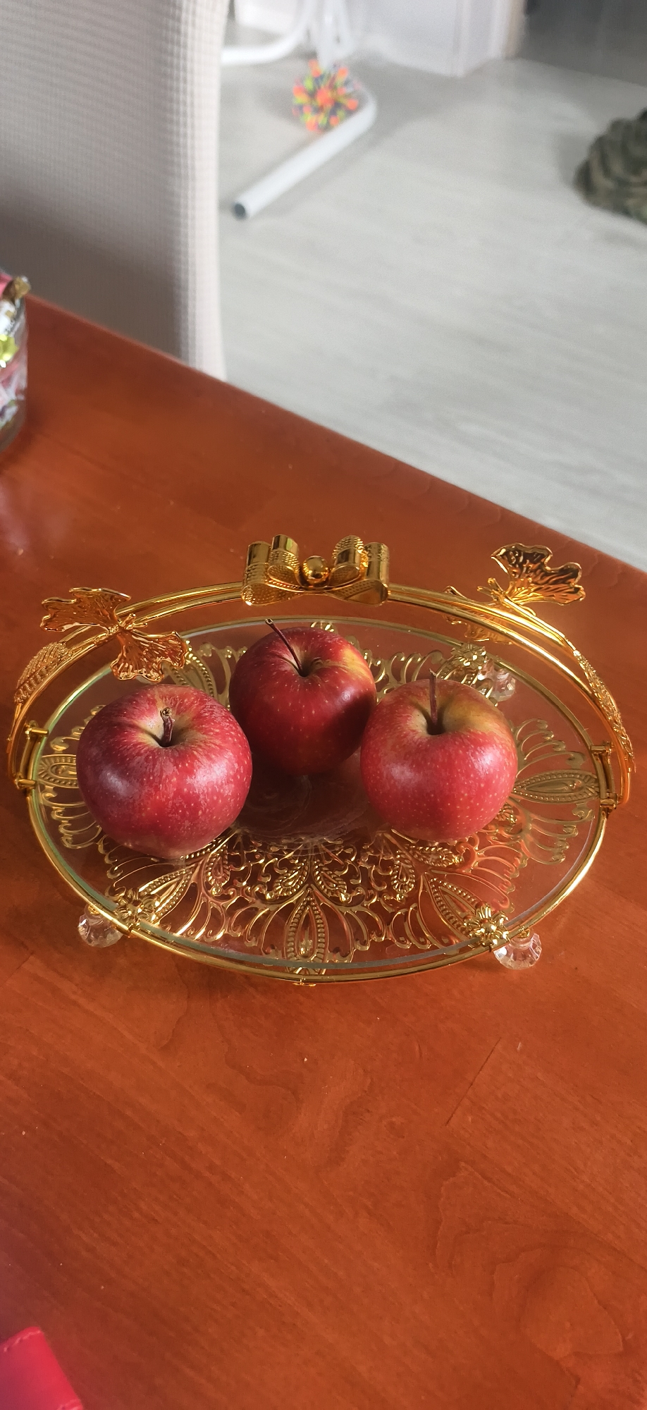 A high-angle, full shot shows three red apples on a gold-trimmed glass tray, placed on a wooden table. The apples are positioned close together in the center of the tray, which is oval-shaped with decorative gold trim and small crystal-like feet. The table has a smooth, polished wood surface, reflecting light. In the blurred background, parts of a chair and a toy are visible. The location is not discernible from the image.