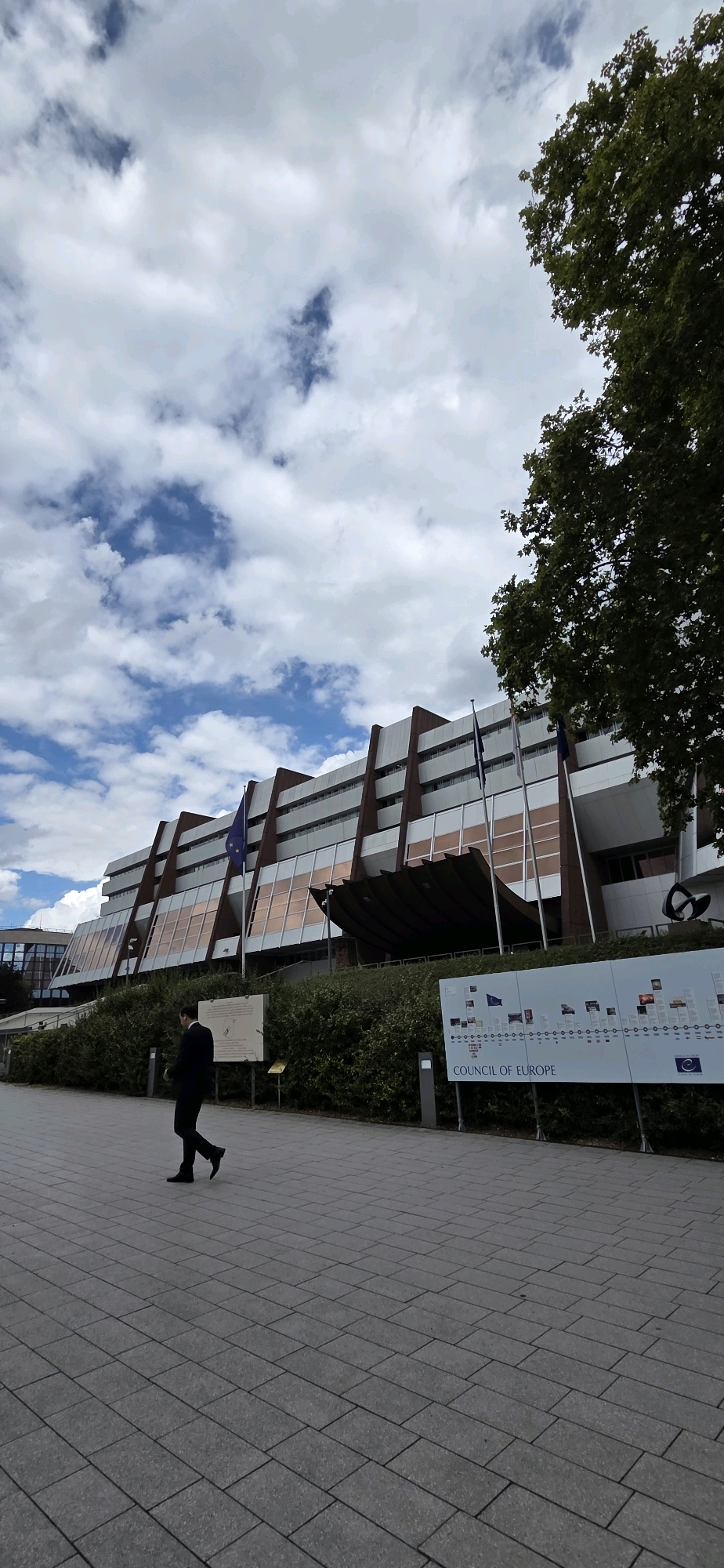 The image shows the Palace of Europe, the seat of the Council of Europe, in Strasbourg, France. A man in a black suit walks on a paved pathway in front of the building. The building is large and modern, with a distinctive angular design and a facade featuring rows of windows and copper-colored panels. Several flags, including a blue flag with stars, fly in front of the building. To the right of the building is a long informational display with the words "COUNCIL OF EUROPE" and a timeline of events. The sky is partly cloudy with patches of blue sky visible. A large tree with green foliage is visible on the right side of the image, partially obscuring the building. The ground is paved with rectangular grey stones.