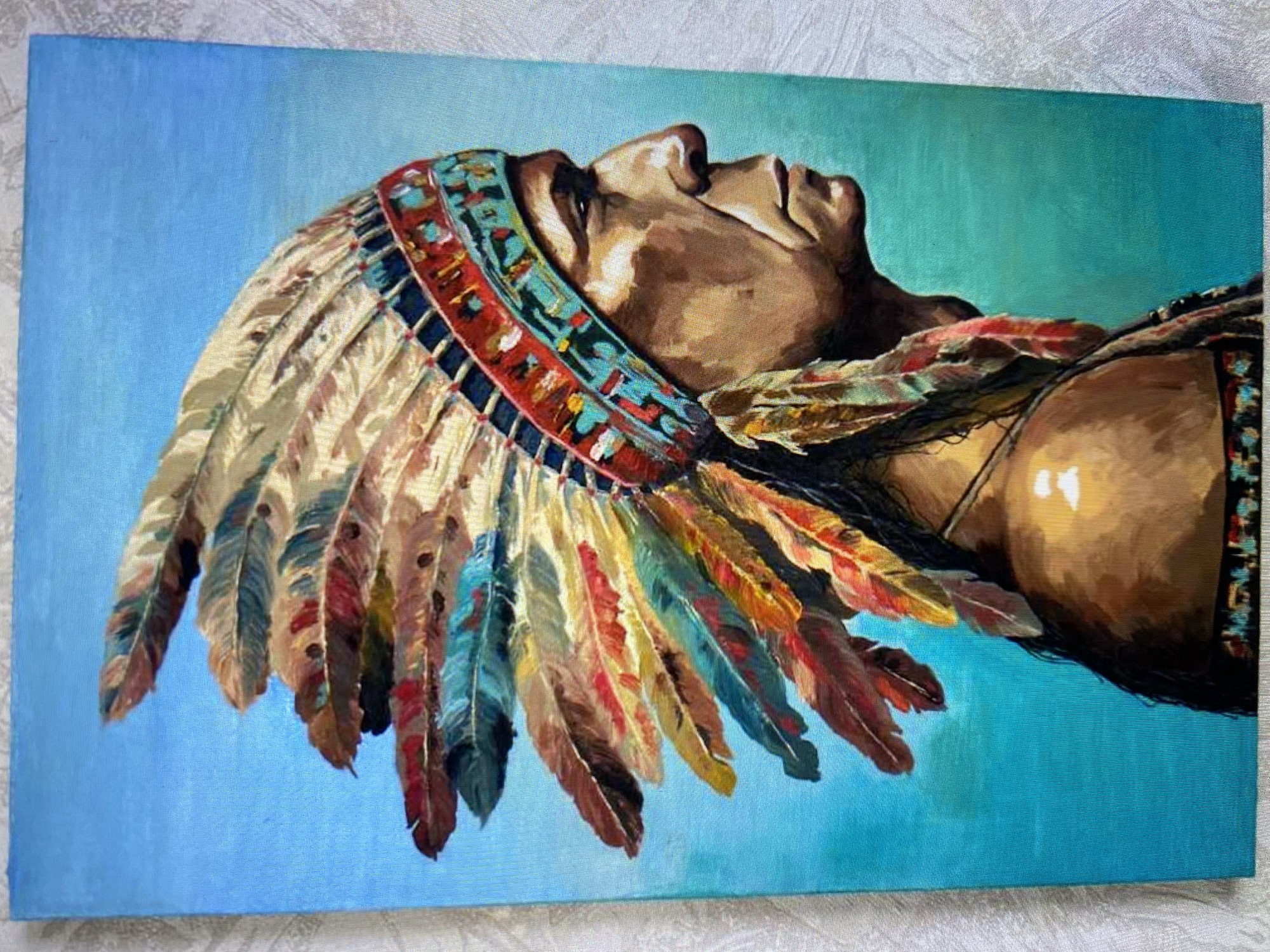 A painting depicts a Native American man in profile, facing right, against a bright blue background. He wears an elaborate feathered headdress with various colors including red, yellow, blue, and brown. The headdress is adorned with a colorful band around his forehead. The man's face is rendered with strong features, and his expression appears stoic. He has dark hair and is wearing traditional attire, including a necklace and possibly beaded clothing. The painting is detailed, with visible brushstrokes, and the lighting highlights the contours of his face and the textures of the feathers. The overall impression is one of dignity and cultural representation.