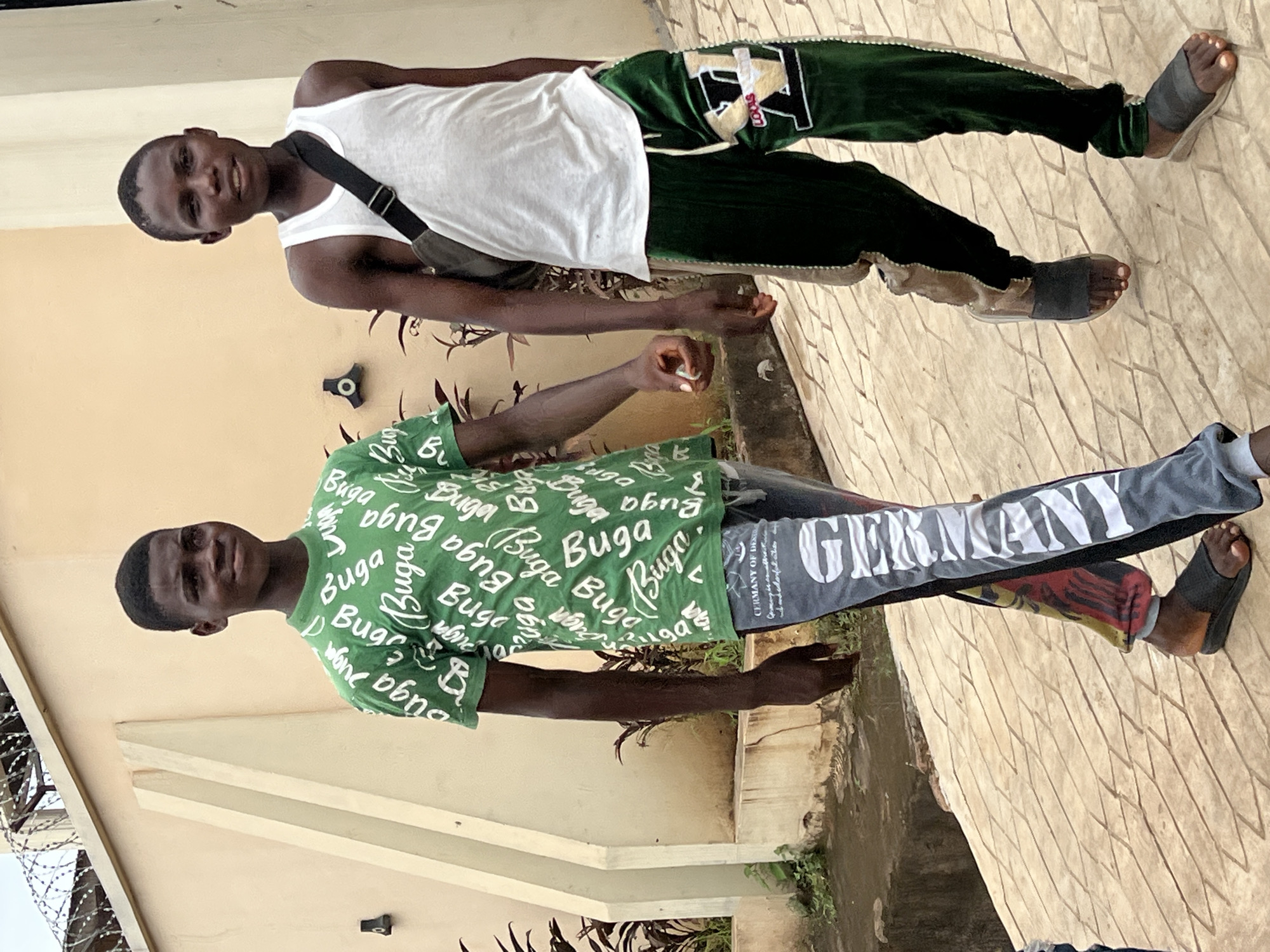 Two young Black males are walking on a paved pathway outdoors. The male on the left is wearing a green t-shirt with white writing and gray and black patterned pants. The male on the right is wearing a white tank top and green velour pants with white trim and a logo on the left leg. He is also carrying a dark-colored bag slung across his chest. The background consists of a light-colored building with angular architectural features and some vegetation. The pathway appears to be made of textured concrete pavers. The sky is visible at the top of the image. The scene is likely located in Nkpor, Nigeria, based on the context provided.