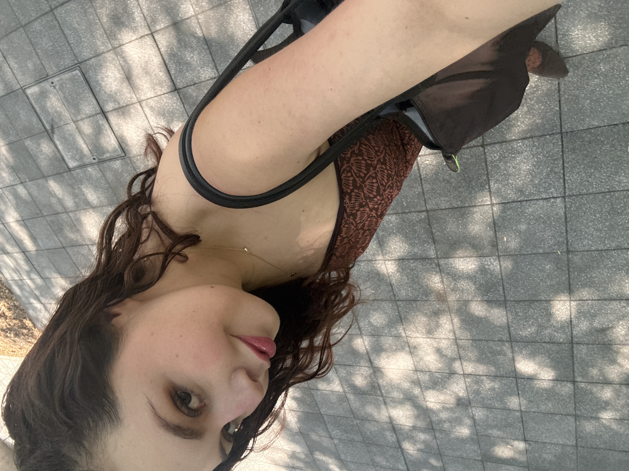 A woman with brown hair, fair skin, and red lipstick is pictured from a low-angle point of view. She is wearing a brown and black sleeveless top with thin straps and a necklace. A black bag hangs on her shoulder. The background consists of a gray tiled surface with shadows of trees. This image was taken outdoors in Mexico City, Mexico.