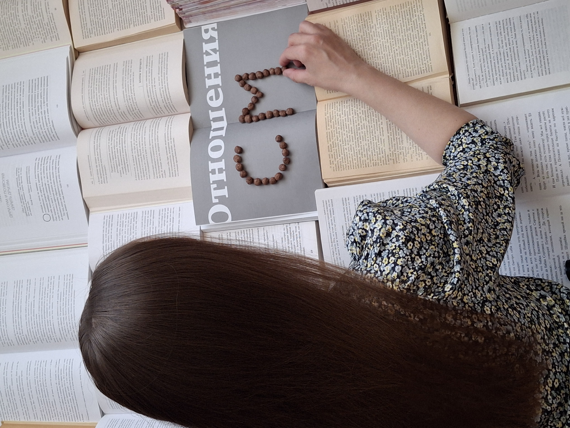 An overhead shot captures a person, likely female, with long, straight, dark brown hair and a floral dress, engaged in a creative activity amidst a spread of open books. The person's right arm and hand are visible, carefully arranging small, brown, spherical objects (resembling chocolate cereal or nuts) on the surface of a prominent open book. These objects are formed into the Cyrillic letters "CMN".
The central book features a large, gray title in Cyrillic, "Отношения" (Otnosheniya), which translates to "Relationships." Surrounding this central book are numerous other open books, all displaying pages filled with dense Cyrillic text, suggesting an academic or literary setting. Visible fragments of text from these pages include phrases like "ДАРИИ" (Darii), "ВОЛНЫ БУДУЩего" (Waves of the Future), "Случай в Бестон" (Case in Beston), and "Арашивает Печ-" (Asks Pech-), indicating Russian language content.
The person is wearing a dark dress adorned with small, intricate floral patterns in yellow, white, and blue. The scene is brightly and evenly lit, suggesting natural daylight. The overall impression is one of focused engagement in a scholarly yet playfully creative environment. The activity of forming letters with edibles on a book titled "Relationships" could imply a personal message or a thematic exploration. The location context for this image is Chaikovsky, Russia.