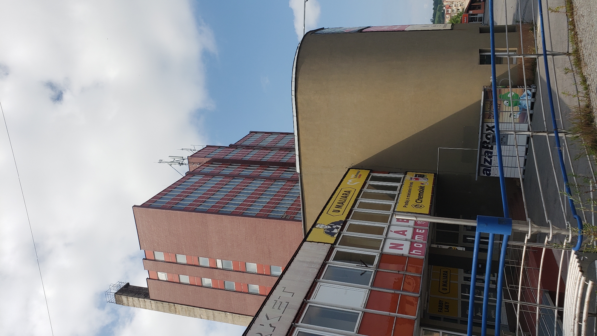 The image depicts a modern commercial building complex on a sunny day in Prešov, Slovakia. In the foreground, a blue metal railing encloses a walkway leading down to an entrance. Adjacent to this is a section of a building with a facade featuring multiple windows and signage. One sign reads "MARKET" in large white letters. Below this, a yellow sign with the text "U MALIARA" and a cartoon illustration of a painter is visible, along with smaller signs that say "NÁPOJE HOME" and "FARBY LAKY".
Behind this lower section, a curved, tan-colored building rises. To the left of this curved building, a tall, older building with a reddish-brown brick facade dominates the background. This building has numerous windows with blue frames and a concrete chimney-like structure extending from its left side. Several cell phone antennas are mounted on the roof of the reddish-brown building.
To the right of the curved building, a modern AlzaBox parcel locker system is installed against the wall. Further in the background, other buildings and a hillside can be seen, suggesting a semi-urban or suburban environment. The sky is partly cloudy with a clear blue sky visible.