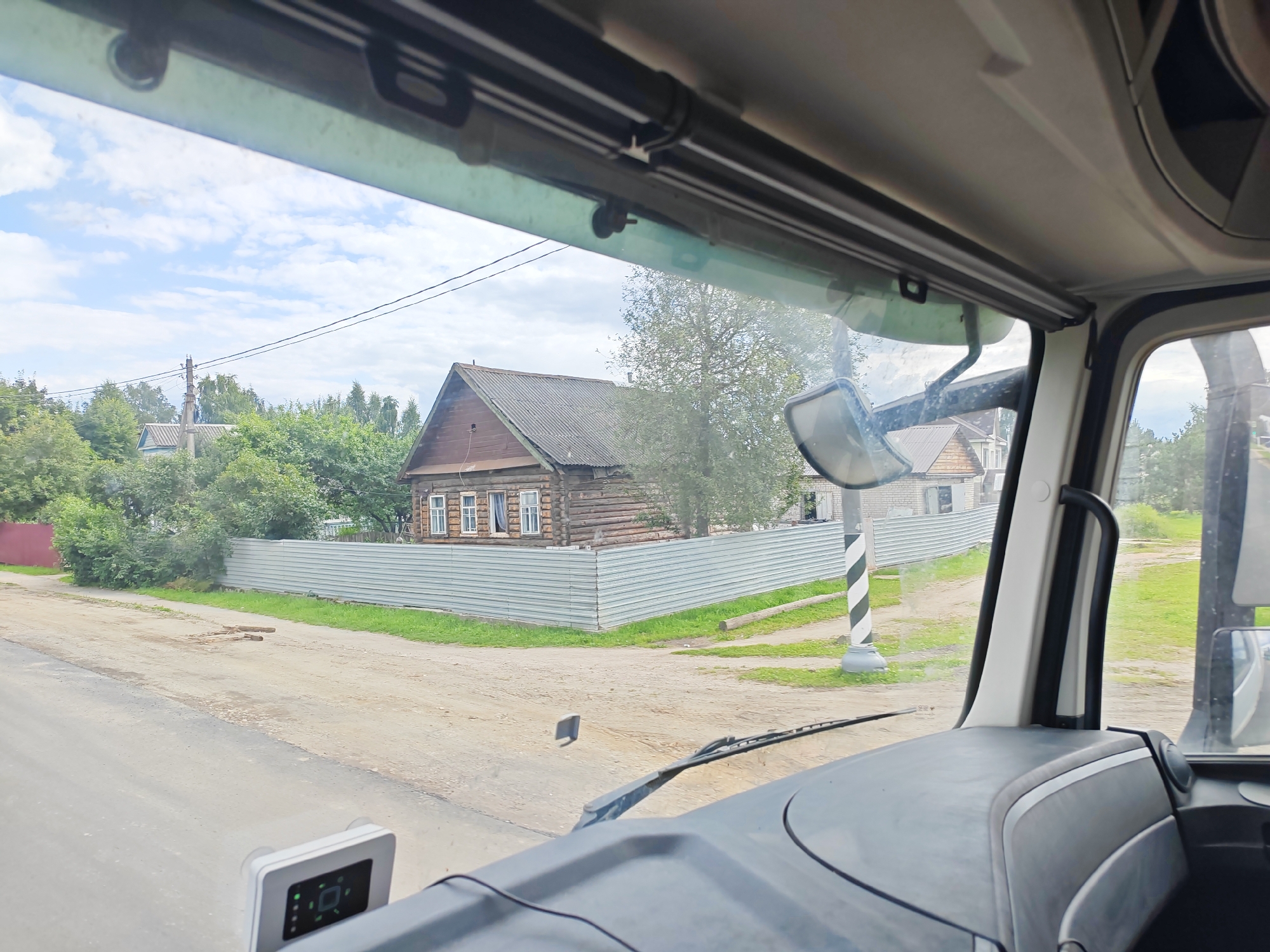 The image is a view from inside a vehicle, looking out the windshield at a rural scene in the village of Выползово, Russia. In the foreground, the dashboard of the vehicle is visible, with a suction-cup mounted device on the lower left. Outside, a dirt road runs parallel to the vehicle. On the right side of the road is a black and white striped pole. Further into the scene, a wooden house with multiple windows is visible behind a light-colored metal fence. Green trees and vegetation are abundant, and power lines are strung across the sky. The sky is partly cloudy.