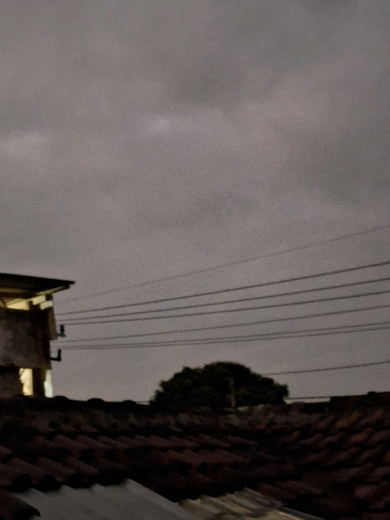 The image shows a nighttime view of a rooftop and the sky in Lumajang, Indonesia. In the foreground, there are tiled rooftops. Behind the rooftops, a silhouette of a tree is visible against the hazy sky. Several power lines stretch across the scene horizontally. On the left side of the image, part of a building with a lit window can be seen. The sky is overcast and dark, suggesting it is either evening or a cloudy day. There are no people or animals present in the image.