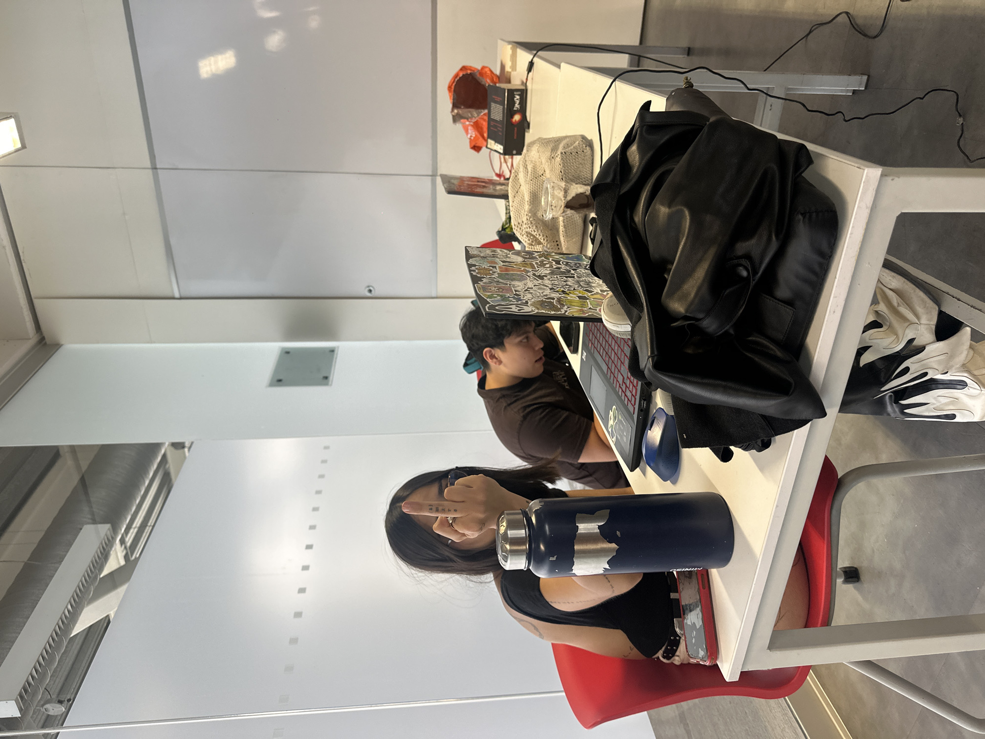 Two individuals are seated at white desks in what appears to be a library or study area. The person closest to the camera is a woman with dark hair, wearing a black tank top and seated in a red chair. She is extending her middle finger towards the camera. A blue water bottle is on the table in front of her, partially obscuring a red smartphone. The other person, a male wearing a brown shirt, is seated further back at another desk with a laptop covered in stickers. A black leather jacket is draped over the desk near him. Other items on the desks include a white crocheted bag, a red box, and black cables. The room has white walls and a frosted glass partition. The environment suggests an indoor academic setting. The city of Mexico City, Mexico is not identifiable within the image's details.