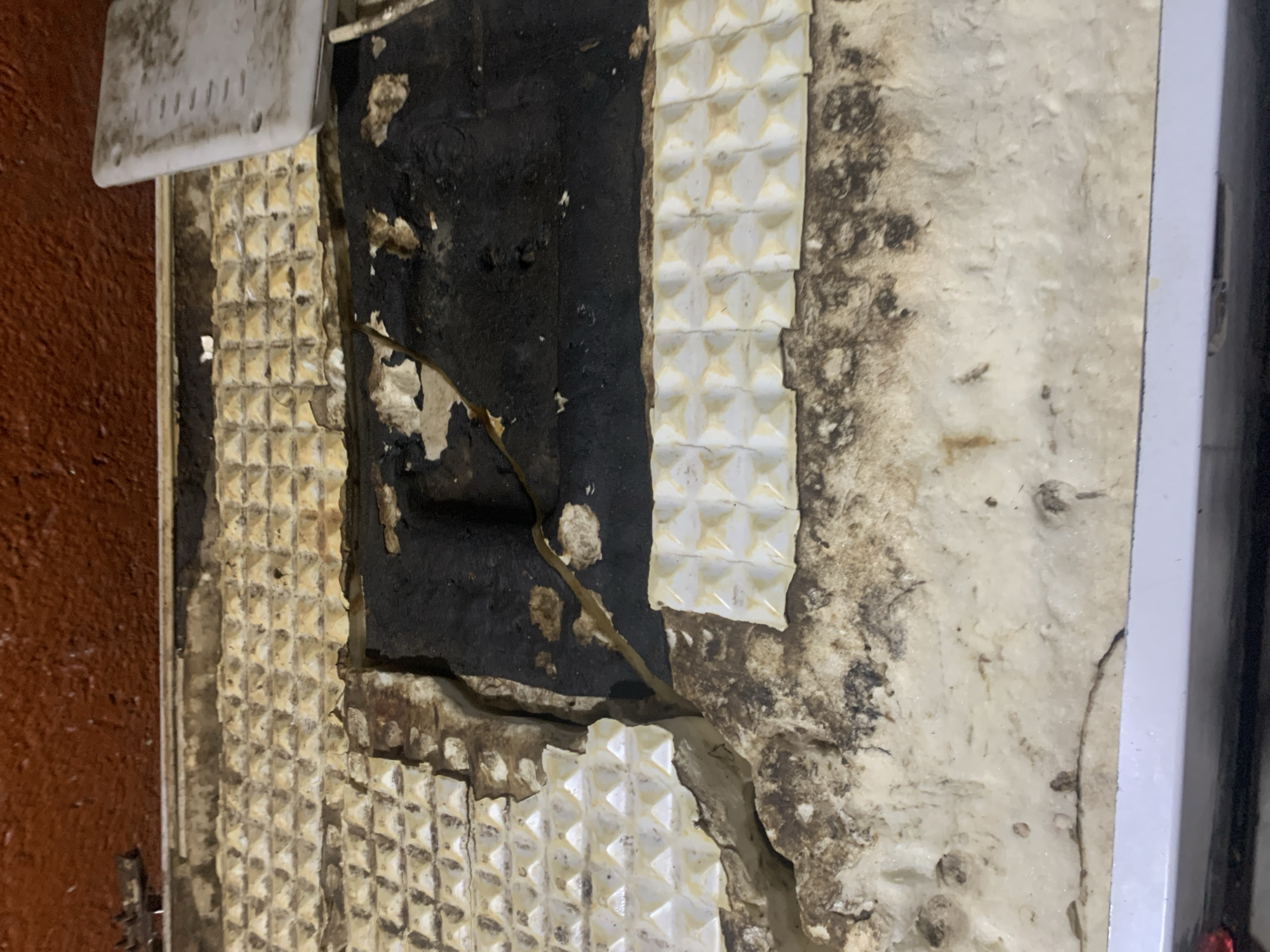 The image shows a close-up, overhead view of a damaged, textured surface, possibly the interior of an appliance or a ceiling. The surface is primarily white with a repeating pyramid-like texture, but it is heavily soiled and deteriorating. Sections of a black, rubbery material are exposed, with some appearing torn or cracked. There are also areas of brown discoloration and what looks like mold or grime. The top edge of the image shows a reddish-brown, textured surface, possibly a wall or ceiling. A light-colored, rectangular object with visible perforations is partially visible on the right side, which might be a cover or panel. The overall condition suggests neglect and possible water damage. The image is not specific to any location within Jos, Nigeria, as it lacks any identifiable landmarks or context.