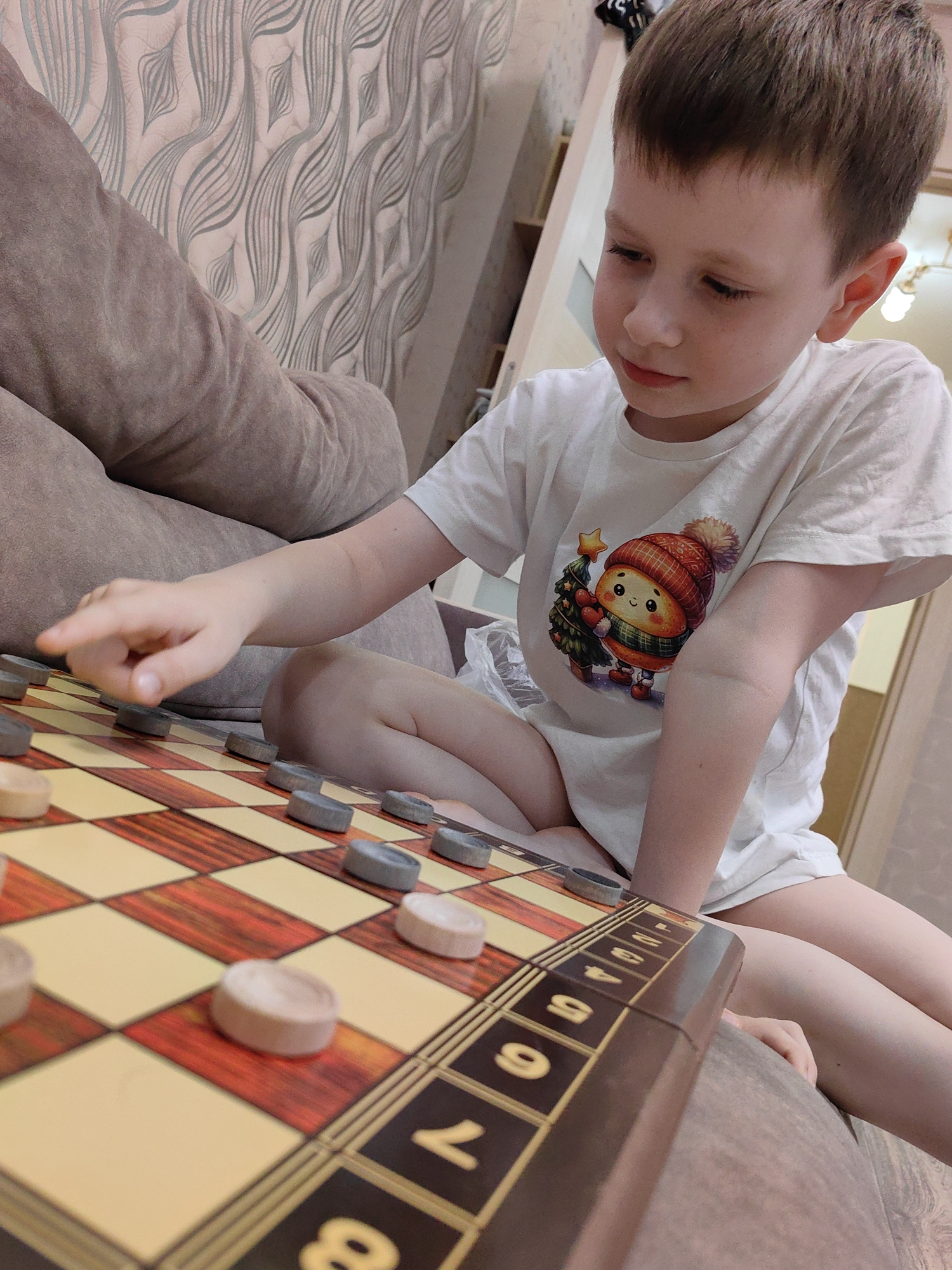 A young boy, appearing to be around 6-8 years old, is sitting on a couch and playing checkers. He is wearing a white t-shirt with a cartoon image of a character holding a Christmas tree. The checkerboard is positioned on his lap, and he is in the process of moving a piece with his right index finger. The background shows a textured wallpaper and a part of a couch. The scene takes place indoors, in what is presumed to be a residential setting. There are no visible indicators in the image to suggest this location is in Sochi, Russia.