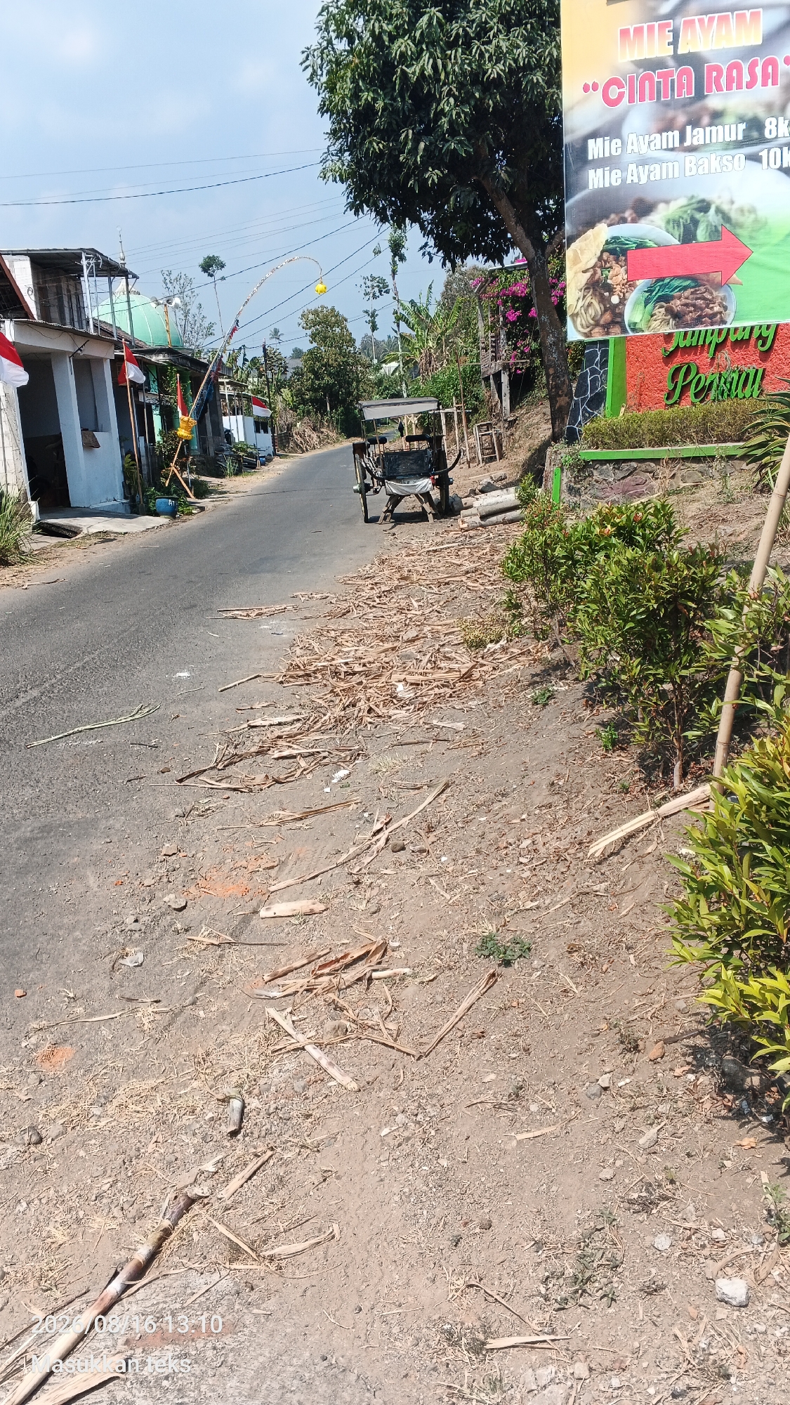 The image shows a street scene in the city of Jeru, Indonesia. The street is paved and appears to be an area with commercial establishments. A sign in the upper right corner advertises "MIE AYAM CINTA RASA" (Chicken Noodles Love Taste), with prices for different types of noodle dishes. Next to the sign is a small, green-roofed building with red and white Indonesian flags displayed. In the middle of the street, a two-wheeled cart with a white canopy is parked. The side of the road is covered with dry, brown debris, possibly dried vegetation or sugarcane stalks. There are also some small green plants growing on the side. The sky is clear and blue, indicating a sunny day. The scene appears to be a typical street in a rural or semi-urban area of Indonesia.