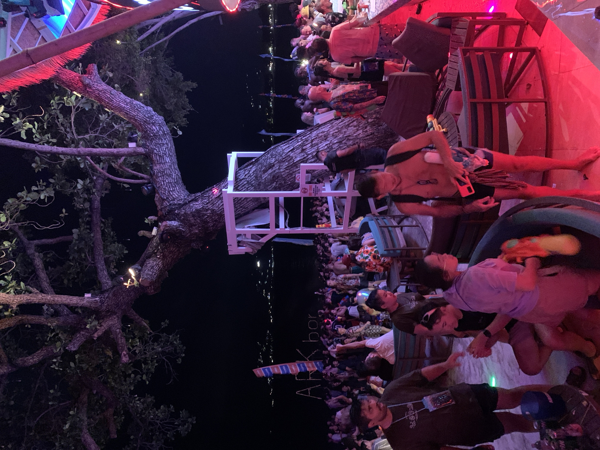 The image depicts a lively scene at a nightspot in Baan Chaweng, Thailand. A large tree dominates the left side, with its branches extending towards the center. A white, platform-like structure is built around the trunk of the tree. Crowds of people are gathered in the background. Nearer to the camera, individuals are holding water guns. Neon lighting is visible on the building structures, with red being a prominent color. The word "ARKbar" is visible, angled from bottom left to top right.