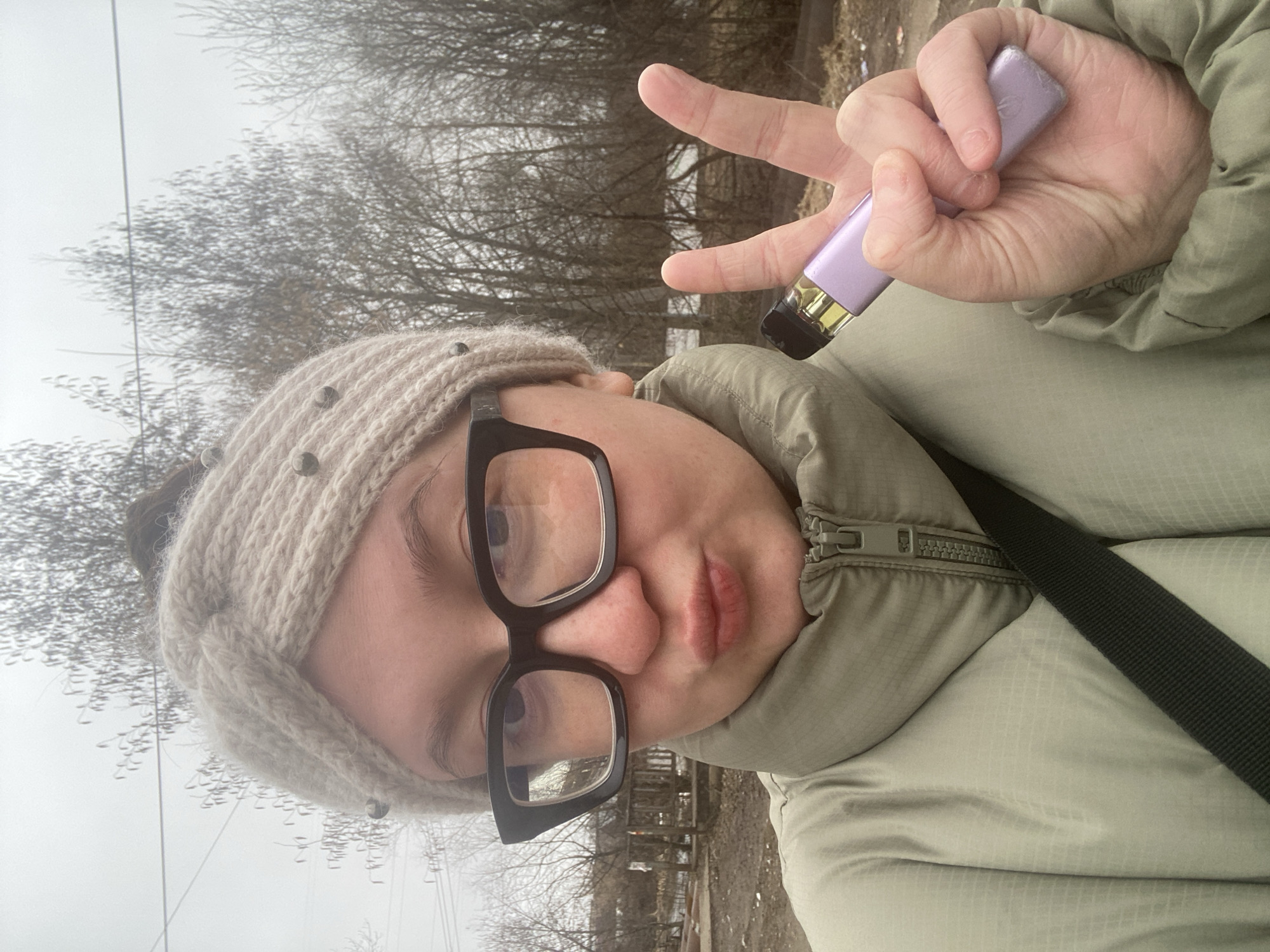 A person is holding a purple disposable vaping device while making a peace sign with their fingers. They are wearing a light green puffer jacket, a light-colored knit headband with studs, and black-framed glasses. The location appears to be outdoors, with bare trees and a somewhat overcast sky in the background. The location is Chaikovsky, Russia.
