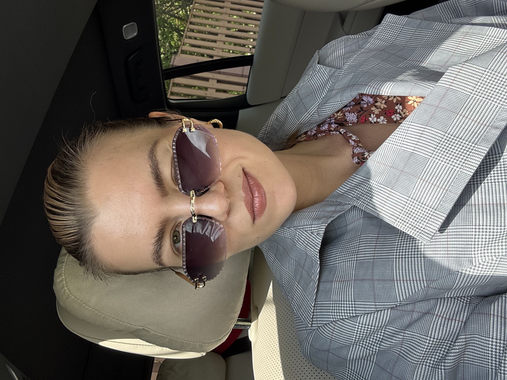 The image shows a woman wearing sunglasses and a patterned jacket sitting in a car. She is angled towards the camera with a slight smile. The car's interior is visible, with a headrest and a glimpse of the car window showing trees outside.