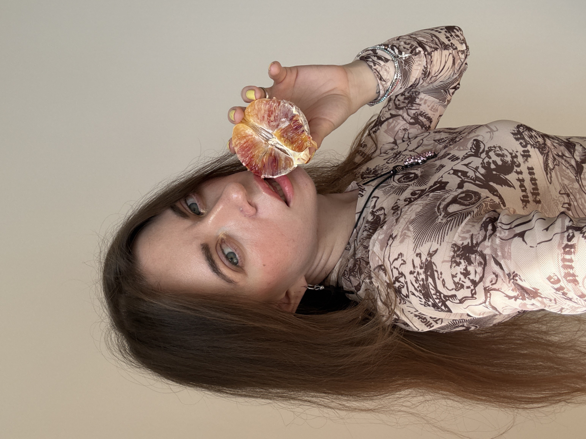 A woman is lying down, holding a halved orange to her lips. She has long brown hair and light blue eyes. She is wearing a patterned, semi-transparent top, and bracelets on her right wrist. She has yellow nail polish. The background is a plain, light beige color. The location is not identifiable as Moscow, Russia, based on the provided image.