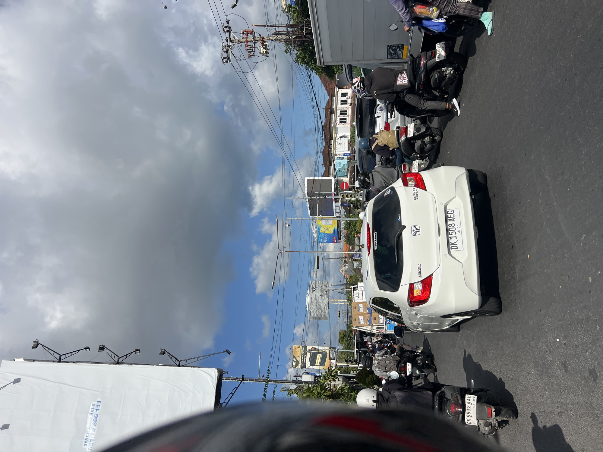 The image depicts a congested street scene in Denpasar Barat, Indonesia, likely during daylight hours. The foreground is dominated by the rear of a white Honda Brio automobile with the license plate "DK 1508 AEG". To the left of the car, a black scooter with the license plate "DK 6812 AAH" is visible. Several other motorcycles, cars, and trucks are present in the traffic jam, indicating heavy road usage. People on motorcycles, wearing helmets and casual attire, are seen navigating the traffic. The urban environment is characterized by overhead power lines, traffic lights, and billboards, one of which displays "SPACE AVAILABLE 031 13 666 111". Buildings with tiled roofs are visible in the background, and the sky is partly cloudy with a mix of blue sky and grey clouds. The road surface appears to be asphalt.