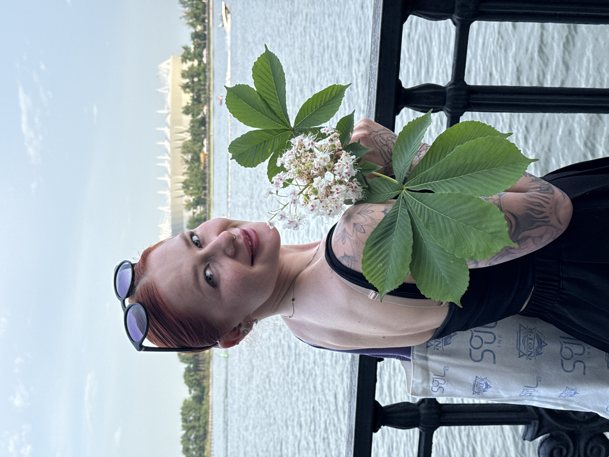 A woman with red hair stands near a railing on a waterfront, holding green leaves and white flowers. Her sunglasses are perched on her head. She has tattoos on her arms. The water and a distant shoreline are visible in the background. Based on the image, it appears to be taken in Rostov-on-Don, Russia, given the tattoo supply company logo is based in the city.