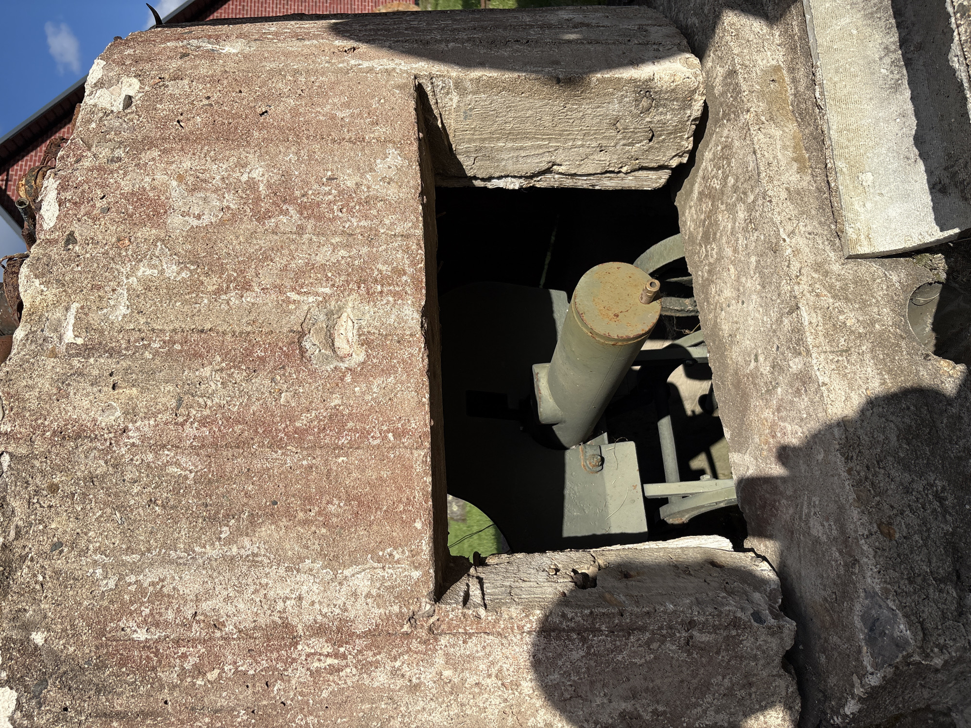 In Zhalybnya, Russia, a weathered concrete structure, possibly a pillbox or bunker, sits partially open, revealing a green, wheeled artillery piece inside. The concrete is rough and discolored with reddish-brown stains, showing signs of age and exposure. The artillery piece appears to be a heavy machine gun or a small cannon, mounted on a metal carriage with two wheels visible. Sunlight casts strong shadows across the scene, highlighting the texture of the concrete and the dark interior of the structure. In the background, a red brick building with a dark roof is visible against a clear blue sky with some clouds. The ground in front of the structure consists of rough concrete and some vegetation.
