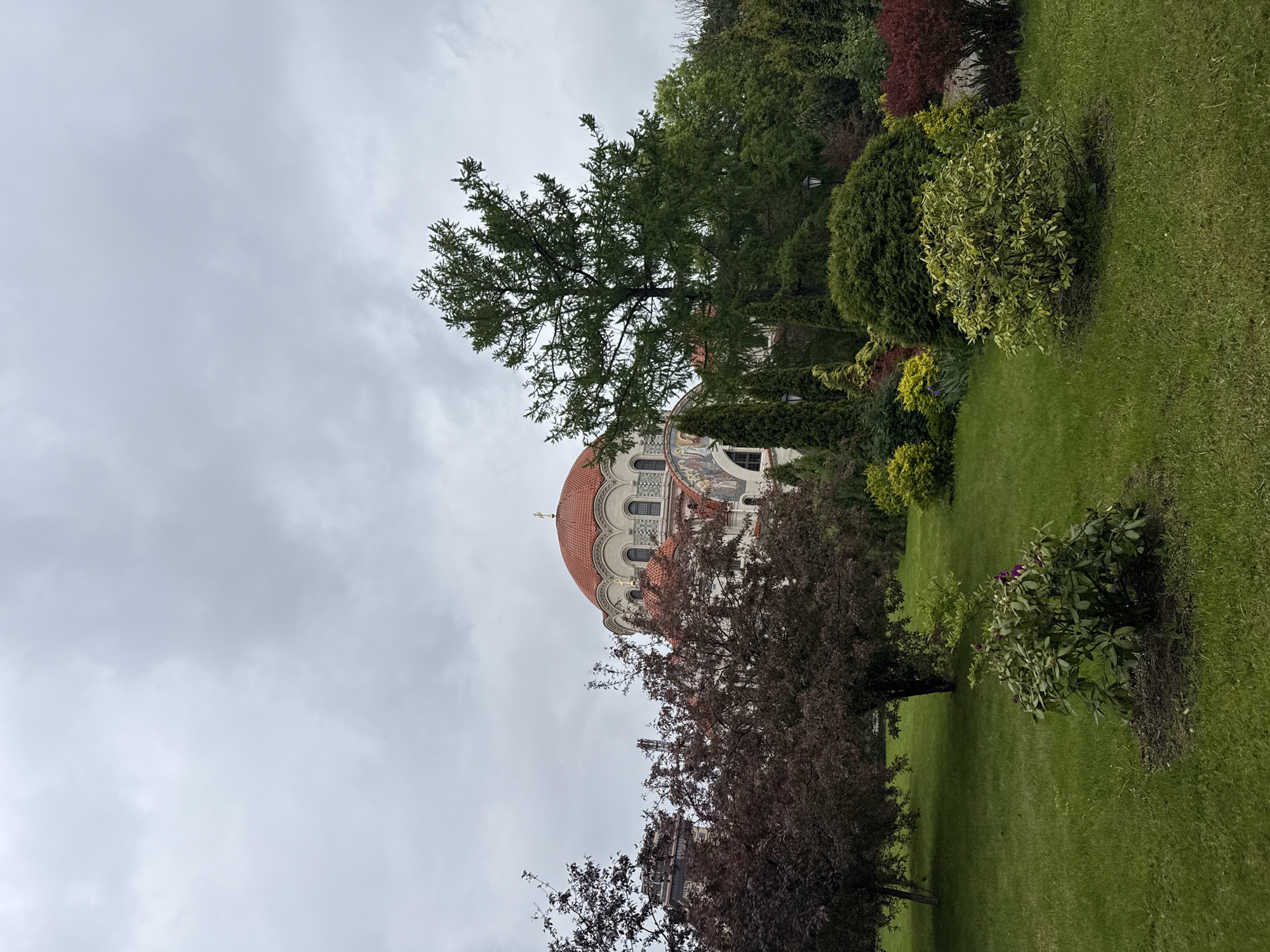 The image shows a partial view of a church with a red-tiled dome, possibly the Church of the Savior on Spilled Blood in Saint Petersburg, Russia. The church is partially obscured by trees and bushes in a grassy area. The sky is overcast and gray. No people are visible in the frame.