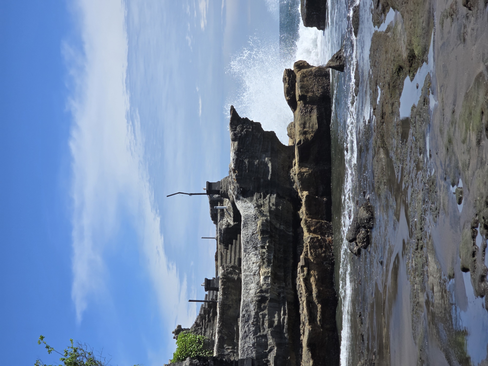 This image presents a dynamic coastal landscape under a bright, clear sky. A prominent, dark, rugged rock formation or sea stack dominates the central and left portions of the frame. This large rock mass appears to have multiple tiers or levels, with what seem to be carved steps or platforms visible along its side, suggesting some form of human accessibility or modification. Several slender, dark posts or poles extend vertically from different sections of the rock formation.
To the right of the main rock, powerful ocean waves are actively crashing against the rocks, generating significant white spray that erupts dramatically into the air. The water at the immediate base of the rocks is turbulent and foamy, while further out, the ocean surface is a deeper blue.
The foreground on the right reveals a dark, wet sandy or rocky shoreline with patches of water, possibly tide pools, reflecting the sky. At the bottom left corner, green foliage from bushes or trees is visible. The sky, occupying the upper portion of the image, is a vibrant blue with scattered, wispy white clouds. The overall scene suggests a sunny daytime setting with clear weather. There is no visible text in the image.