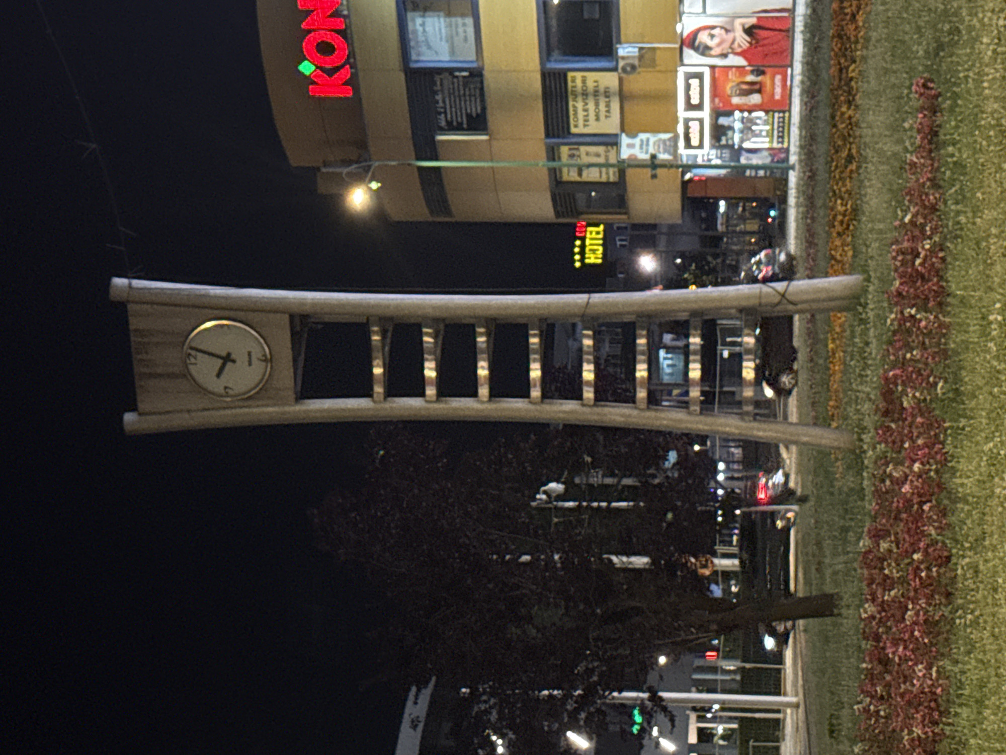 This image captures a street scene at night in Ilidža, Bosnia and Herzegovina. The focal point is a tall, modern clock tower with a light-colored frame and a circular clock face displaying the time as approximately 12:10. To the right of the clock tower is a multi-story yellow building with illuminated signage. One prominent sign reads "KONGRESNI" in large red letters. Below this, other signs are visible, including one that says "HOTEL". The building also has windows displaying advertisements, and an air conditioning unit is visible on the upper facade. In the foreground, there is a well-maintained grassy area with flowerbeds featuring red and yellow foliage. Several cars are parked on the street or driving by. A dark-colored car with its headlights on is visible on the left side of the image, and another car is partially visible on the right. Trees with dark foliage are scattered throughout the scene. The overall atmosphere is that of a quiet urban night, with artificial lighting from the buildings and streetlights illuminating the surroundings.