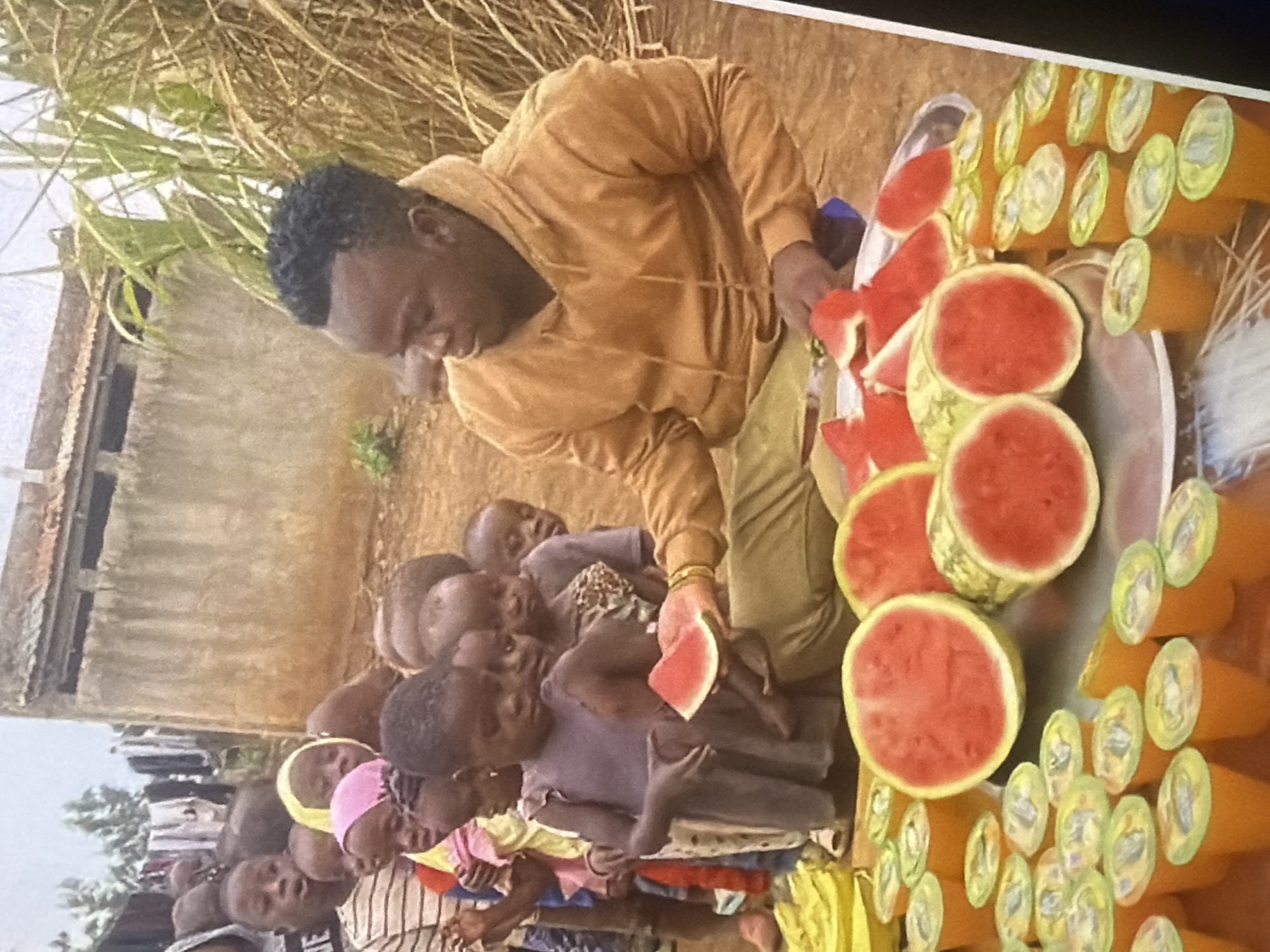 In an outdoor setting, likely a village, a man in a mustard-colored hooded sweatshirt is seated, offering slices of watermelon to a line of children. The children, varying in age, are gathered closely together, some with hopeful expressions as they anticipate receiving the fruit. The environment appears somewhat rural, with a dirt ground and a building with a corrugated roof in the background, possibly a community structure or dwelling. Some vegetation, including tall reeds, is visible to the right of the man. In the foreground, numerous small orange cups with yellow lids are arranged, suggesting they might contain a beverage to accompany the watermelon. The scene conveys an act of sharing and community engagement.