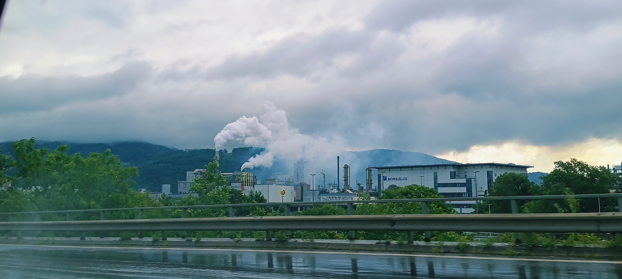 The image captures an industrial landscape in Linz, Austria, on a cloudy day. In the foreground, a wet road reflects the overcast sky and the metal railing of an overpass. Beyond the railing, a lush green tree line partially obscures a complex of industrial buildings. Prominently, a factory chimney emits a large plume of white smoke into the cloudy sky. Various industrial structures, including pipes and smaller buildings, are visible within the complex. To the right, a modern building with the text "BOREALIS" is situated. In the background, rolling, tree-covered hills rise under the diffused light of the cloudy sky. The overall scene suggests an active industrial site located in proximity to a natural, hilly landscape.