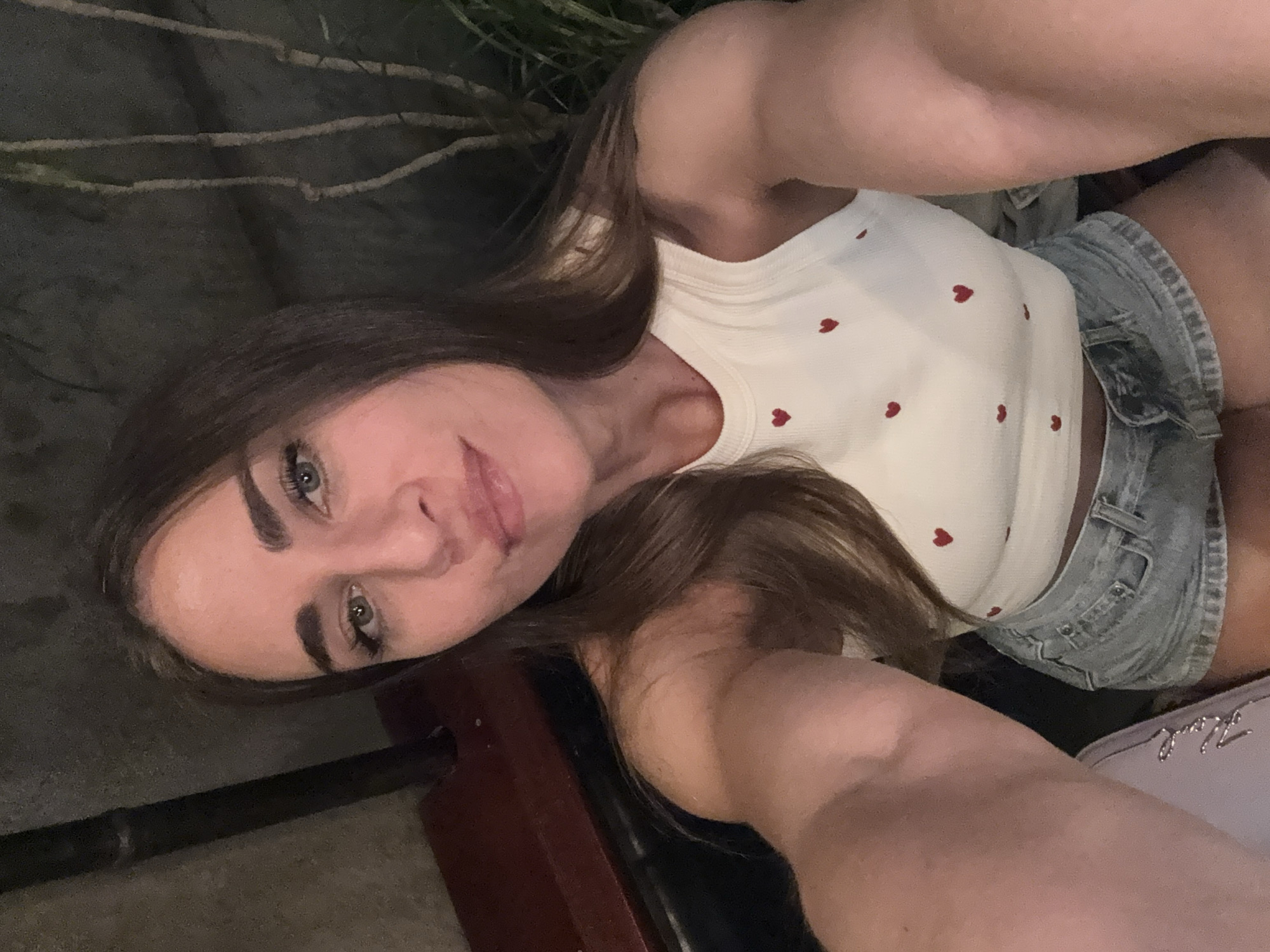 A young woman with long, brown hair is taking a selfie. She is wearing a white tank top with small red hearts printed on it and denim shorts. Her left arm is extended, holding the camera. The background appears to be outdoors at night, with dark, shadowy foliage and what looks like branches. A wooden bench or table is partially visible in the foreground. Her expression is neutral, with a slight smile. The overall lighting is dim, suggesting an evening setting. There is no visible text in the image.