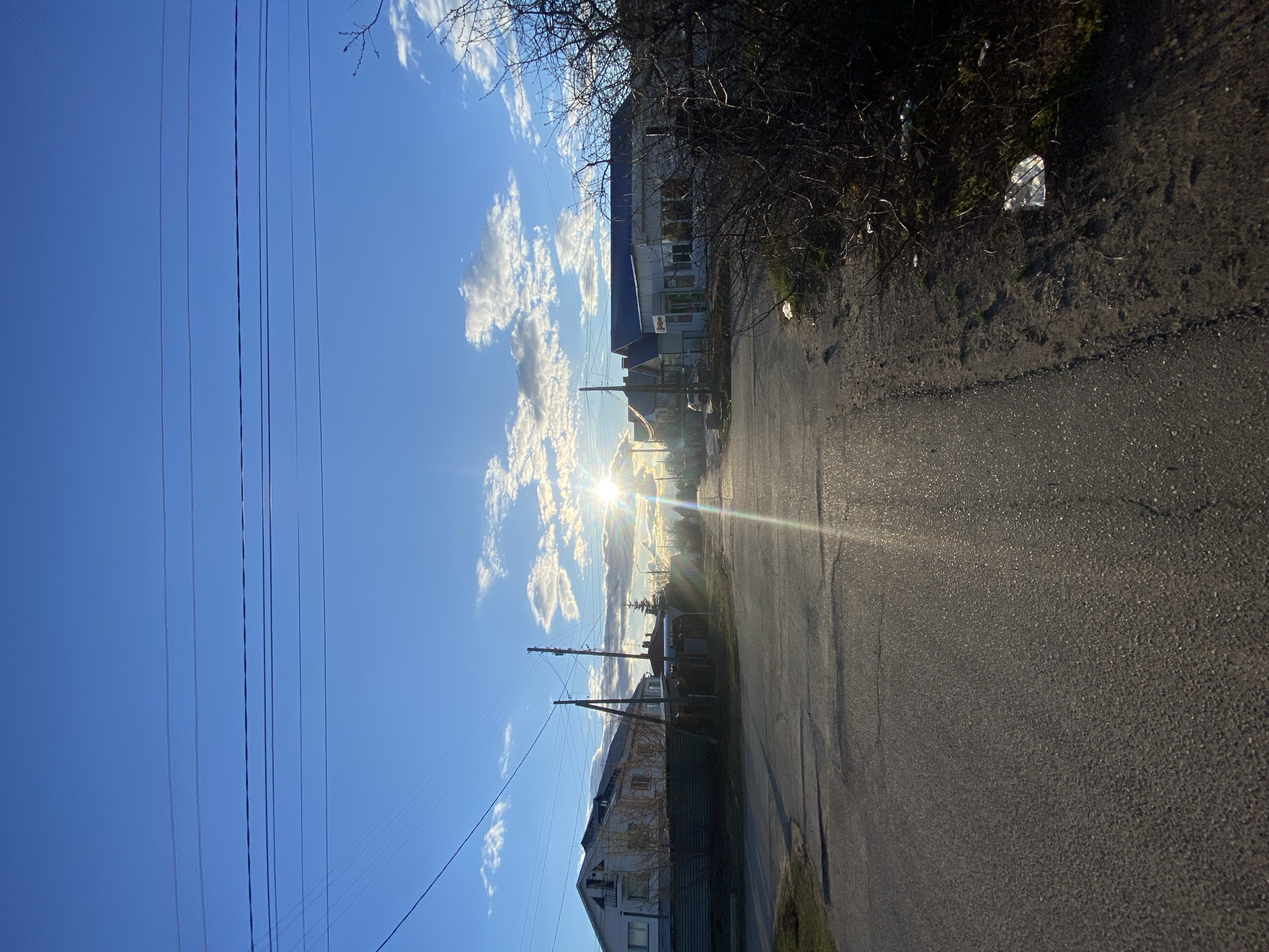 An outdoor scene features a street or path under a clear blue sky with scattered white clouds. The sun is low on the horizon towards the right, casting a bright sunburst and lens flare that illuminates the environment.
Numerous parallel utility wires stretch horizontally across the upper left portion of the image, supported by poles that are faintly visible. Along the right side of the frame, bare tree branches are prominent, suggesting a dormant season. An asphalt road or path extends from the foreground into the distance on the left.
Several buildings line the right side of the road in the mid-ground. One building has a white lower story and a dark upper section or roof. Further down the road, another larger structure with a dark roof and lighter walls is visible. In the lower right corner, beside the road, there is some discarded white plastic trash among dry grass and dirt.
The low angle of the sun indicates either early morning or late afternoon. The weather appears clear and sunny. No people or specific activities are depicted. No legible text is visible on any of the buildings or objects.