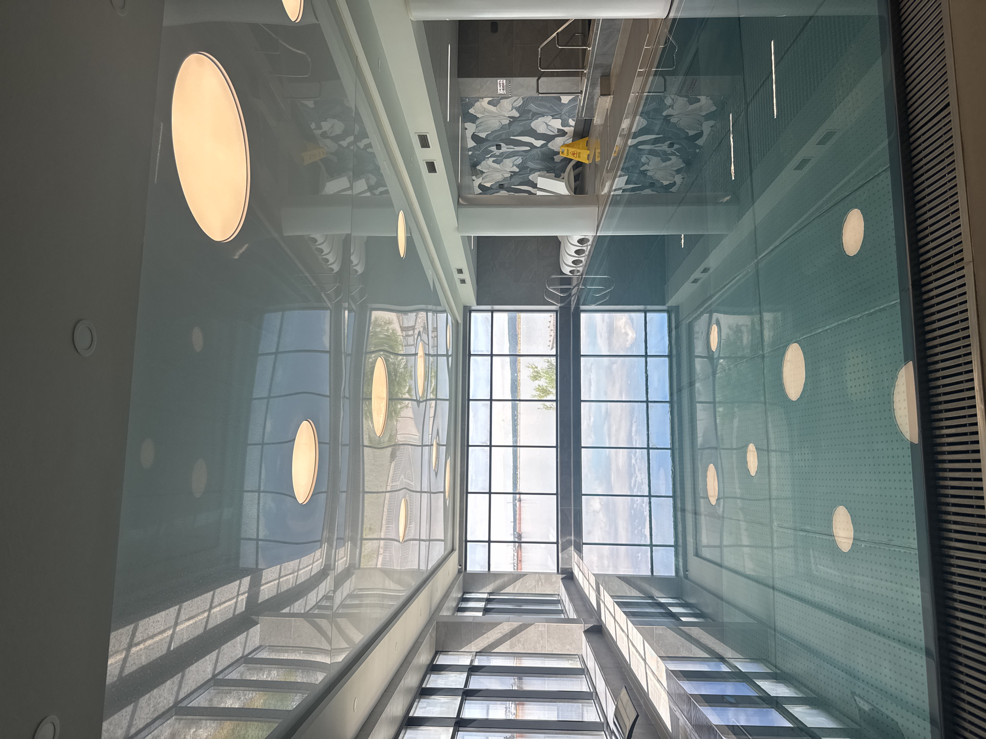 The image depicts an interior view of a building in Chaikovsky, Russia. The location appears to be an indoor swimming pool facility. Large windows show an exterior view of a body of water. The ceiling has circular light fixtures. A patterned ceiling area with a yellow "Caution Wet Floor" sign is also visible. The overall scene is one of modern architecture with natural light integration. No people are directly visible.