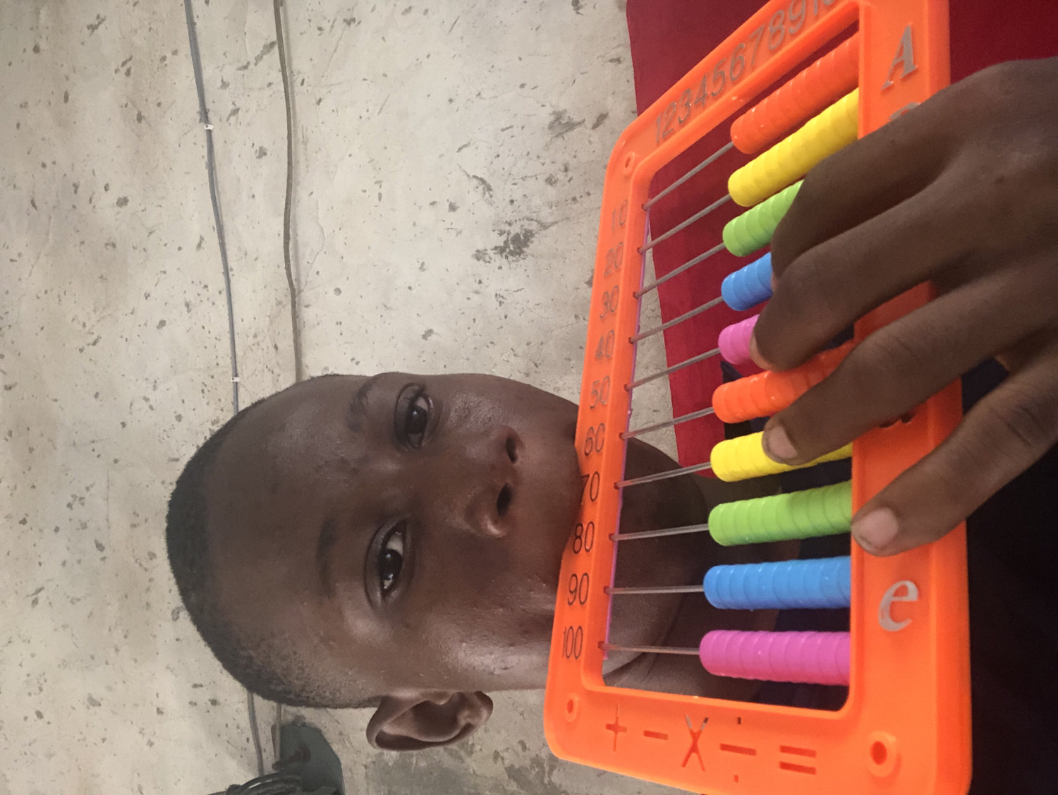 A young person, likely a child or young adult, with dark skin and short, closely cropped hair, is featured in a close-up shot. Their head is tilted, and their gaze is directed towards the camera. The individual's face shows a slightly shiny or sweaty appearance.
They are holding a bright orange plastic abacus-like counting toy with both hands. The abacus features ten horizontal metal rods, each holding ten cylindrical beads in alternating colors of orange, yellow, green, blue, and pink. The right hand's fingers are positioned on the beads, suggesting active use or manipulation of the toy.
Visible text and numbers on the abacus include "123456789" along the top bar and the letter "A" on the top right corner. Along the left vertical frame, numbers "10, 20, 30, 40, 50, 60, 70, 80, 90, 100" are printed. The bottom frame displays arithmetic symbols: plus (+), minus (-), multiply (x), divide (÷), and equals (=). A lowercase "e" is also present on the right vertical frame.
The background consists of a light-colored, possibly concrete or plaster wall with a textured, speckled appearance. Two dark, thin lines, resembling cables or wires, run vertically along the wall. A glimpse of red fabric is visible behind the abacus and the person's hand. The setting appears to be indoors.