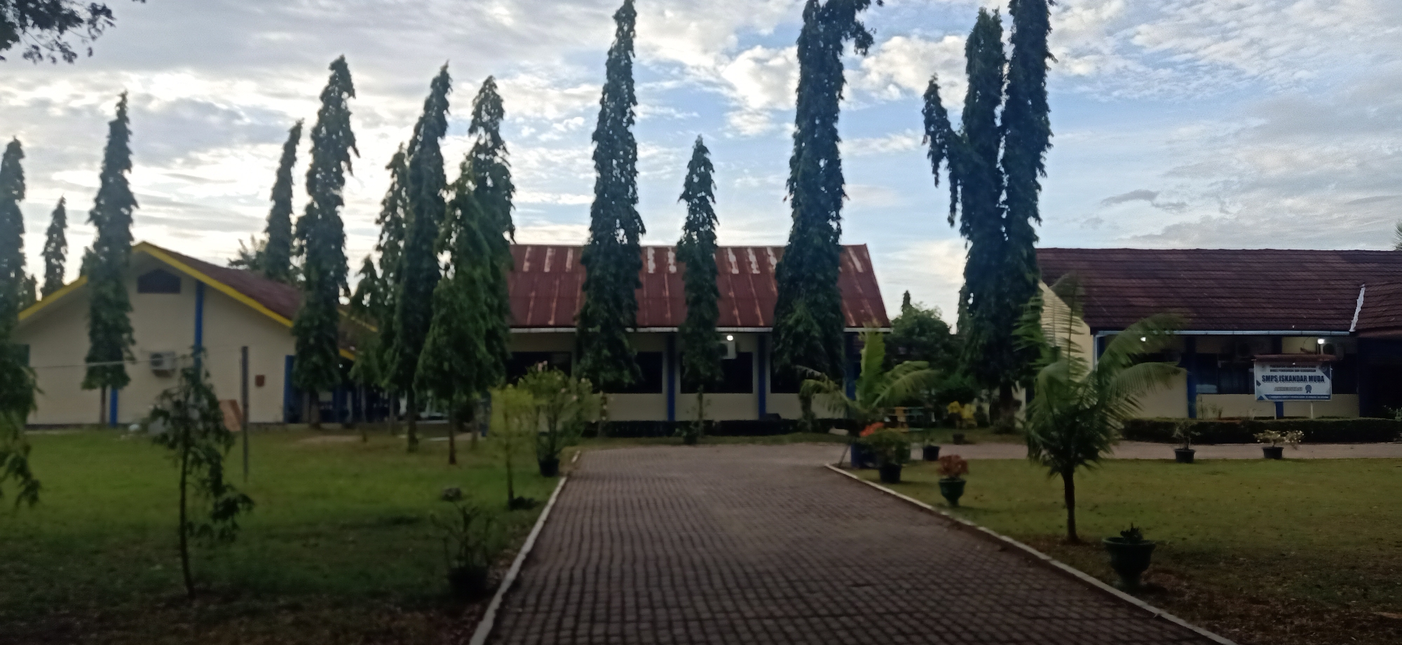 The image depicts a building complex in Tambontunong, Indonesia, likely a school, with a paved walkway leading towards it. The buildings have reddish-brown tiled roofs and light-colored walls, with blue accents. Several tall, slender evergreen trees are planted in rows along the sides of the walkway and in front of the buildings, creating a symmetrical avenue. The ground is covered with green grass, and some potted plants are visible. A sign with Indonesian text, possibly identifying the institution, is mounted on the facade of one of the buildings. The sky is overcast with scattered clouds. The scene appears to be captured during the daytime, possibly early morning or late afternoon, given the diffused lighting. There are no people visible in the image.
