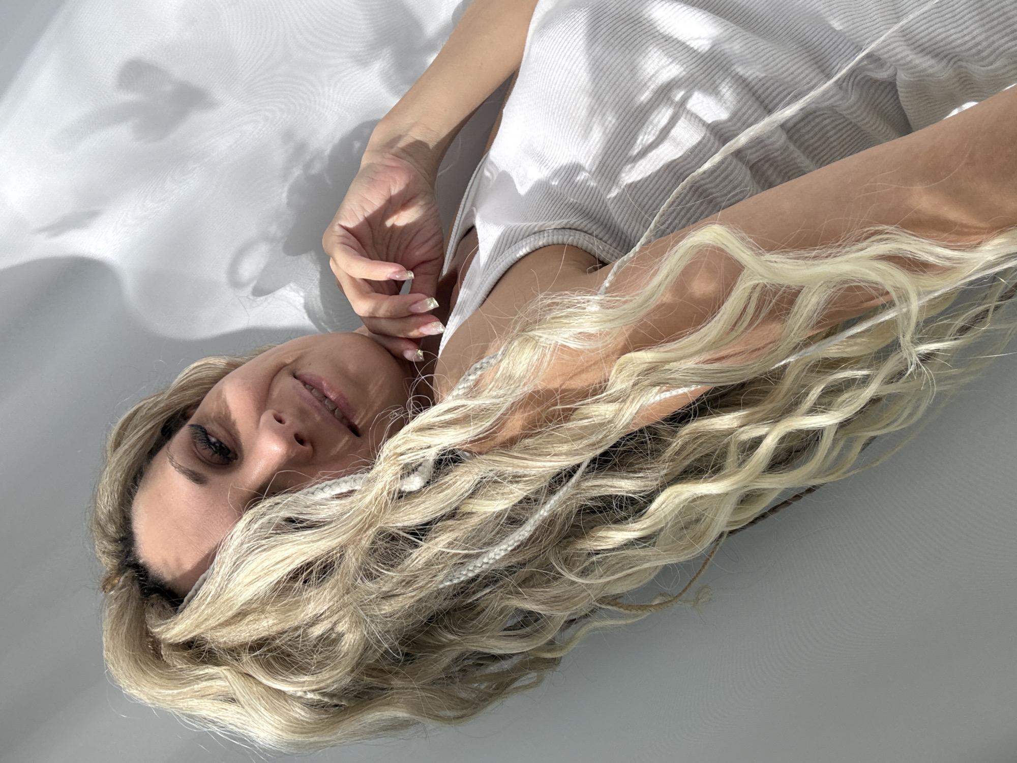 A woman lies on a white surface, her head at the bottom left. She has long, blonde, wavy hair with braids. Her right hand is near her face, fingers slightly curled. She wears a white tank top. Sunlight casts shadows on the wall behind her. The location is not discernible as Rusanovo, Russia based on the visual content of this image.