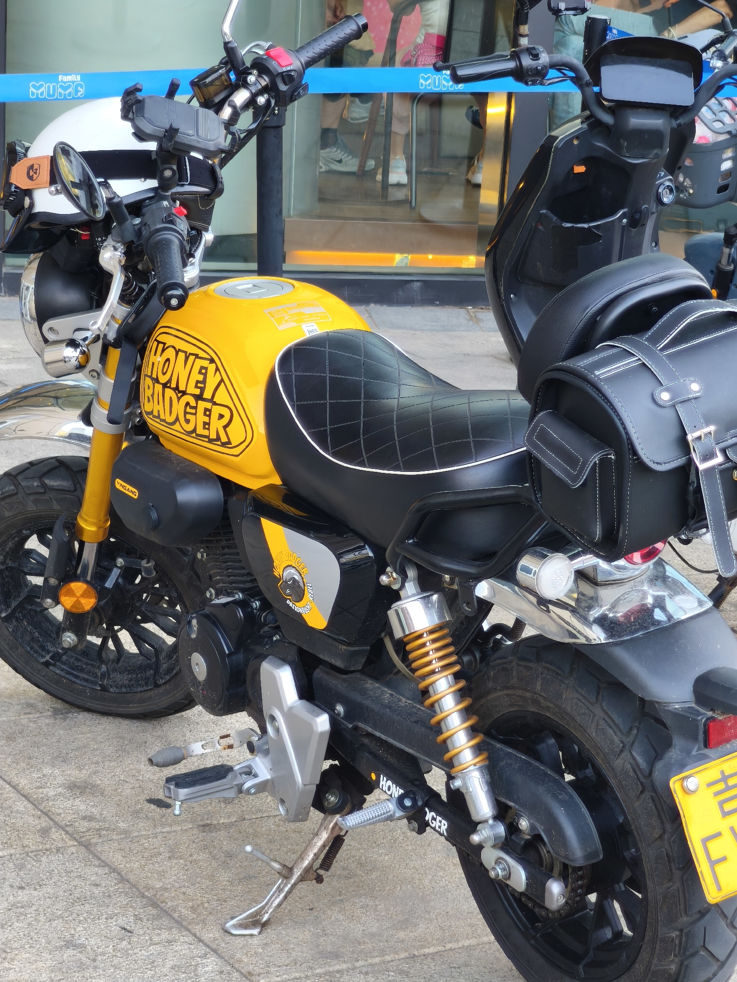 A yellow and black Honda motorcycle with a "HONEY BADGER" decal is parked on a paved sidewalk in front of a store with large glass windows. The motorcycle has a white helmet with a black stripe on the handlebars. A black leather saddlebag is attached to the rear of the motorcycle. The store background shows people walking inside. The location is Changchun City, China.