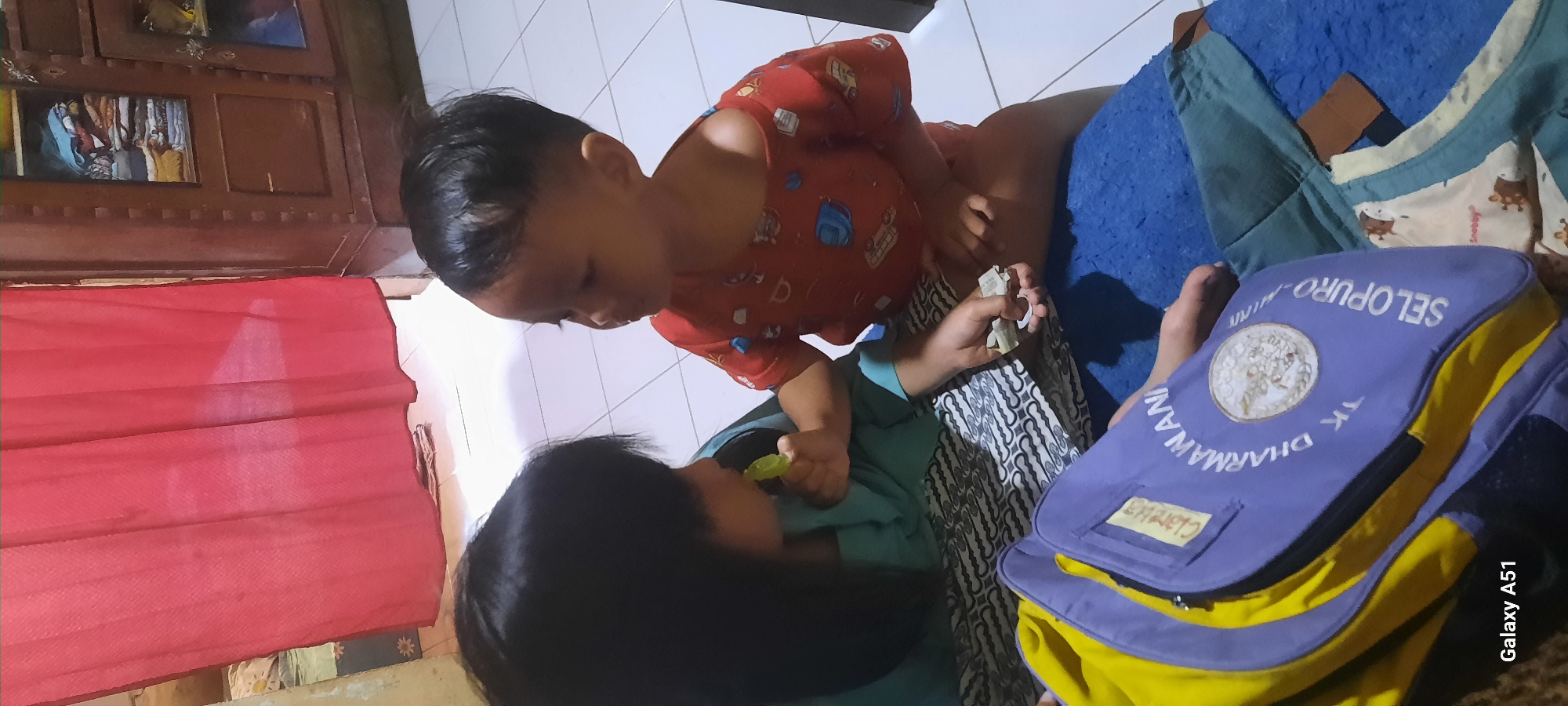 In an indoor setting, likely a residential room in Ploso, Indonesia, two young children are interacting. On the left, a girl with dark hair is wearing a teal top and is in the process of putting a small, green object into her mouth. Facing her on the right is a younger boy wearing a red t-shirt with a pattern of various objects. He is looking at the girl and holding a small, folded piece of paper or receipt in his right hand.
In the foreground, a backpack is partially visible. It is primarily purple with yellow accents and has text printed on it, including "CLARISSA," "DHARMAWAN," "TK SELOPURO," and a circular emblem. The children are sitting on a patterned floor covering, which appears to be batik. The background shows a red curtain on the left and a wooden cabinet filled with folded clothes on the right. The floor is tiled with white squares.