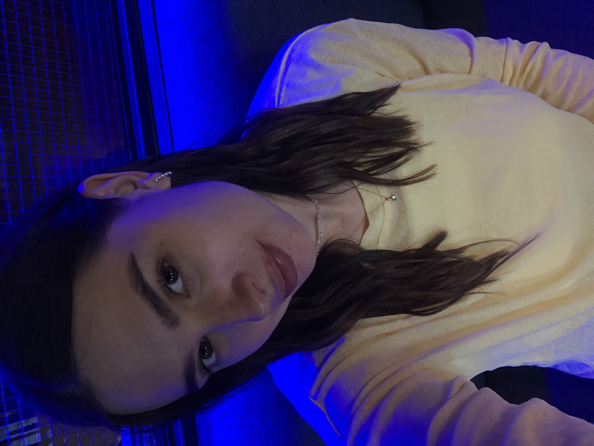 A young woman with dark brown hair and fair skin is taking a selfie. She is wearing a yellow long-sleeved shirt and a delicate necklace with a small pendant. Her makeup includes lipstick and mascara. She has a small stud earring in her left ear. The background is dark, with a prominent blue neon light creating a blue cast on her hair and shoulder. There is a mesh-like structure in the upper background, also illuminated by the blue light. The scene appears to be indoors, possibly in a bar or club, given the lighting and background elements. There is no discernible location in Izhevsk, Russia, or any other specific city visible or implied in the image.