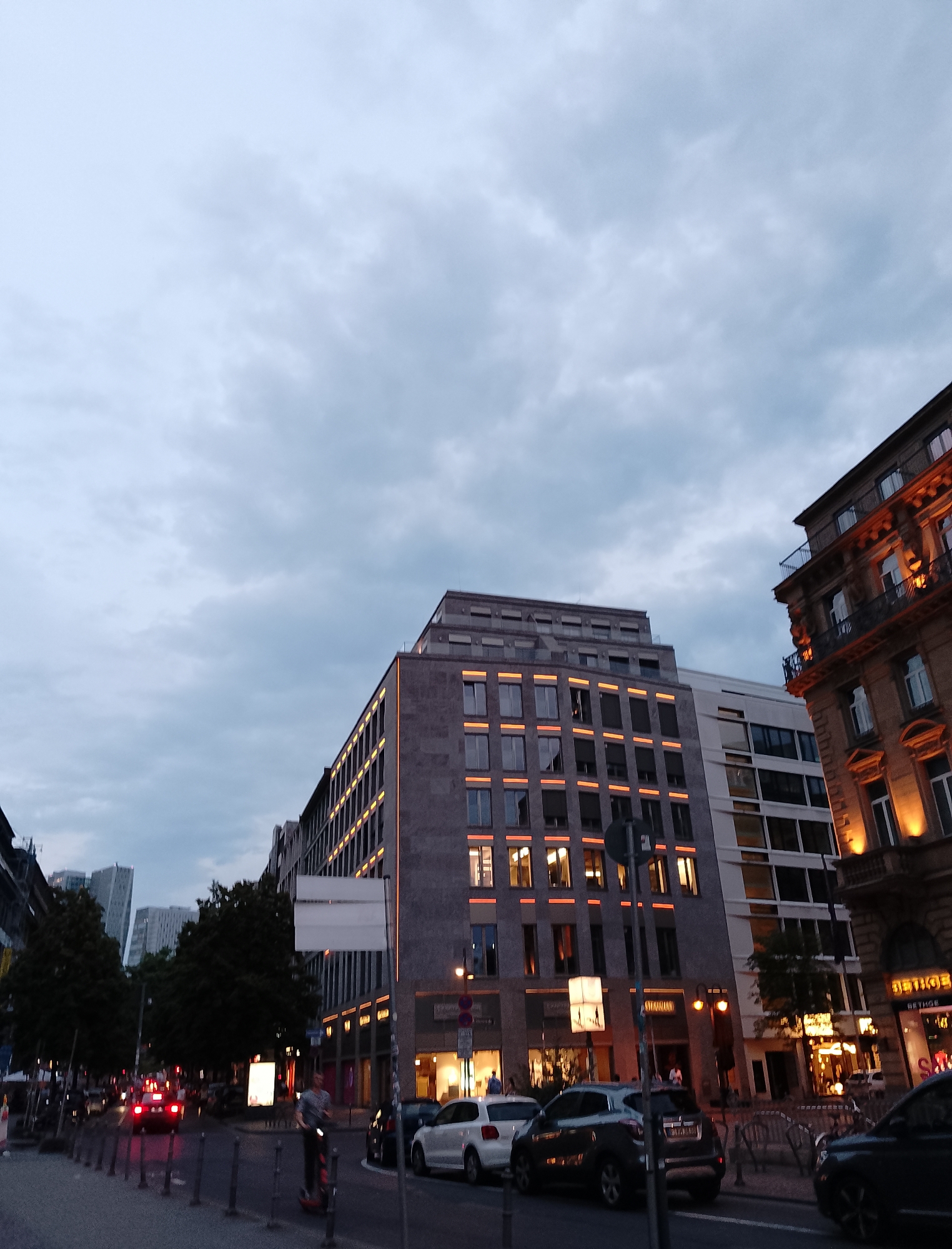 The image shows a street scene in a city at dusk or early evening. The sky is overcast with clouds. On the right side of the image, there is an ornate building with balconies and illuminated windows. To the left of this building is a modern building with a gray facade and numerous windows illuminated with orange lighting. Further to the left, another modern building is visible, and in the distance, several taller, more contemporary skyscrapers can be seen. The street is paved and lined with parked cars, some of which have their headlights on. A person is riding an electric scooter in the foreground, and another person is walking on the sidewalk. Trees are present along the street. The scene suggests an urban environment with a mix of architectural styles.